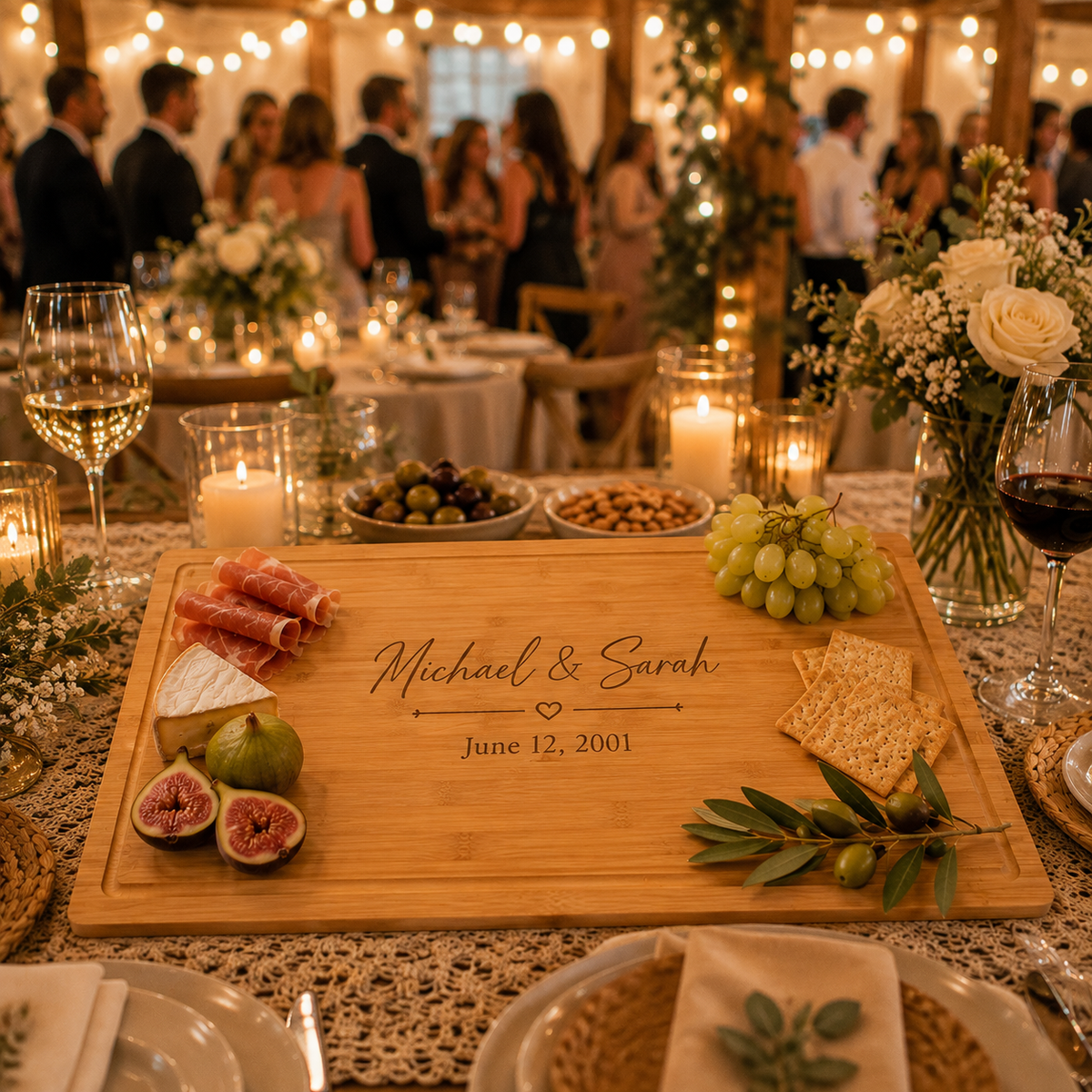A personalized wooden charcuterie board engraved with 'Michael & Sarah' and a date, styled on a decorated wedding reception table with cheese, grapes, crackers, olives, and candles in the background.