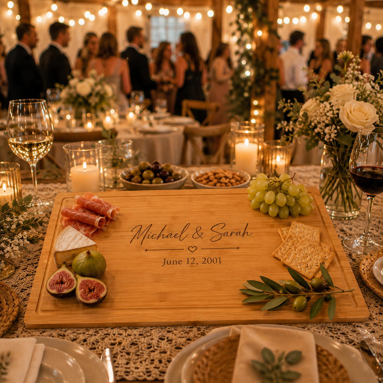 A personalized wooden charcuterie board engraved with 'Michael & Sarah' and a date, styled on a decorated wedding reception table with cheese, grapes, crackers, olives, and candles in the background.