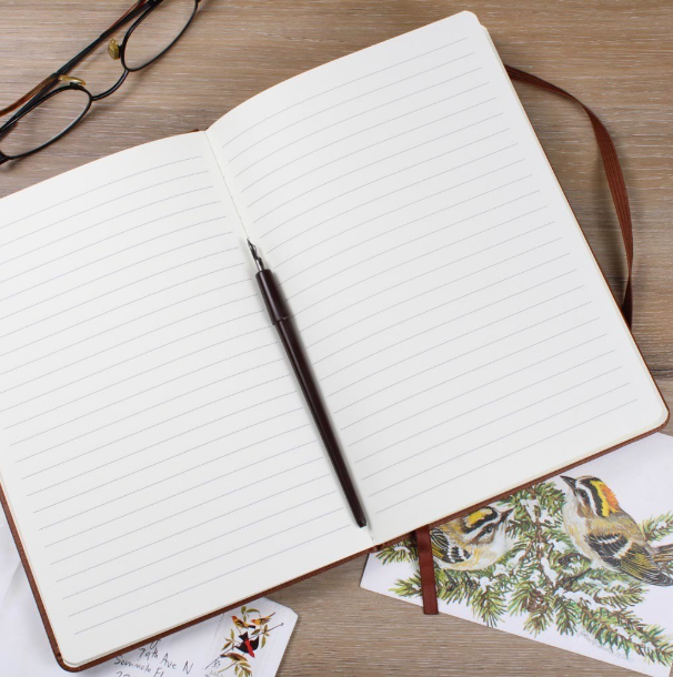 Open notebook with lined pages and brown pen resting on right side, vegan leather cover with burgundy binding visible, styled on wooden surface with eyeglasses and botanical illustrations nearby