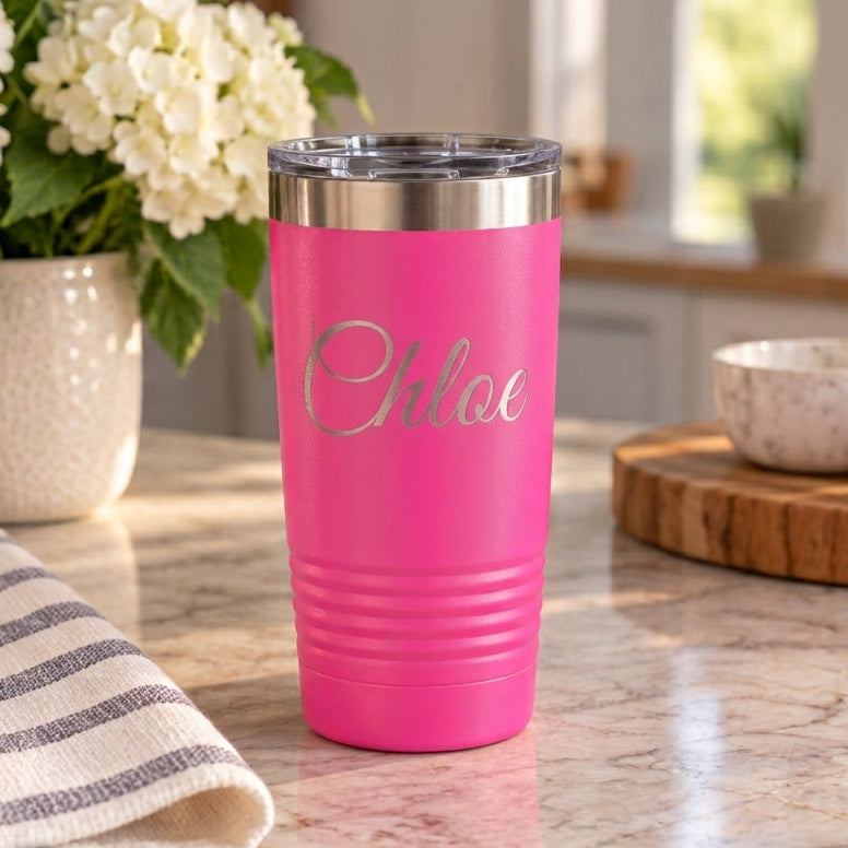 A bright pink insulated tumbler with a clear lid and the name 'Chloe' printed in silver script on the front, set on a marble kitchen counter with flowers and bowls in the background.