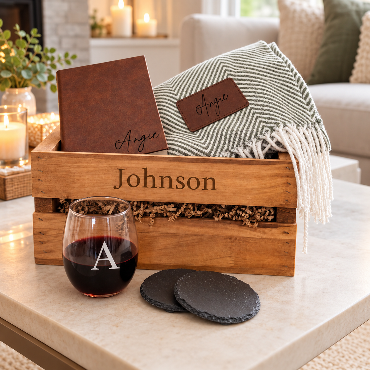 a crate with the name Johnson on it with a stemless wine glass with a A monogram etched on it, 2 slate coasters, a leatherette brown journal, and a blanket grey and white. The background has a couch, pillow and candles