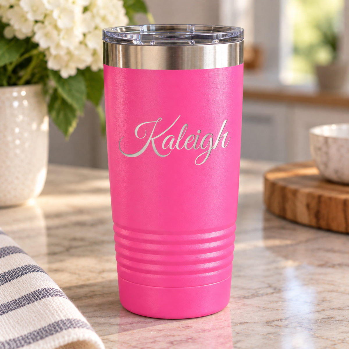 A bright pink insulated tumbler with a clear lid and the name 'Kaleigh' in silver script, standing on a kitchen countertop with flowers and kitchen items blurred in the background.