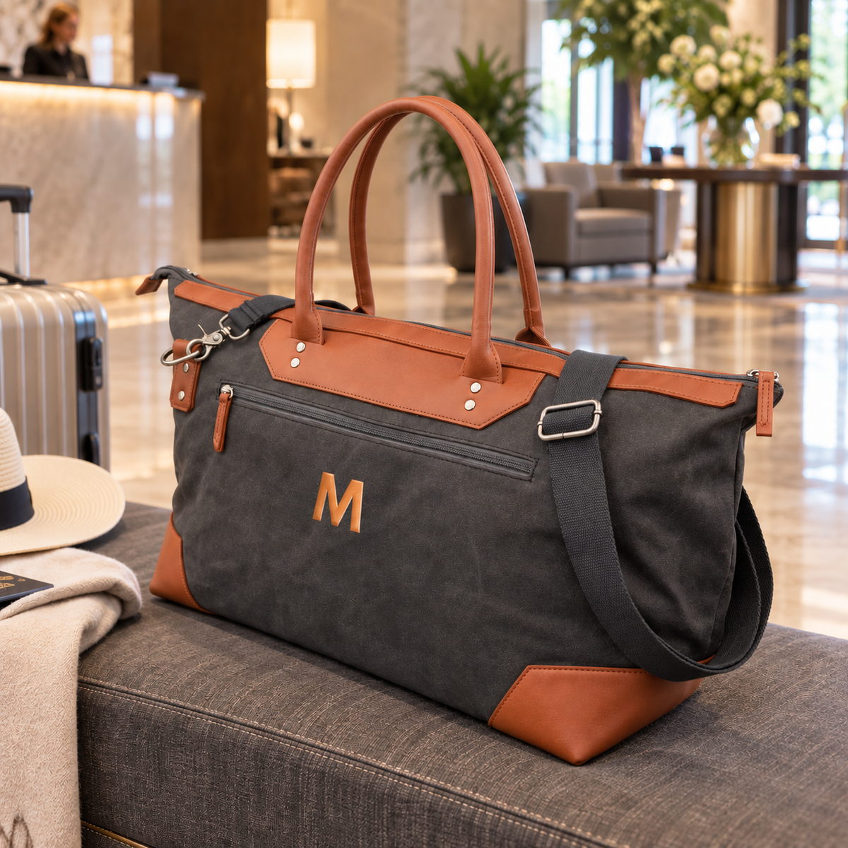 A dark gray canvas and brown leather duffel bag with a monogrammed 'M' sits on a gray bench in a modern, upscale hotel lobby. A striped suitcase, a white hat, and a beige throw blanket are partially visible next to it.