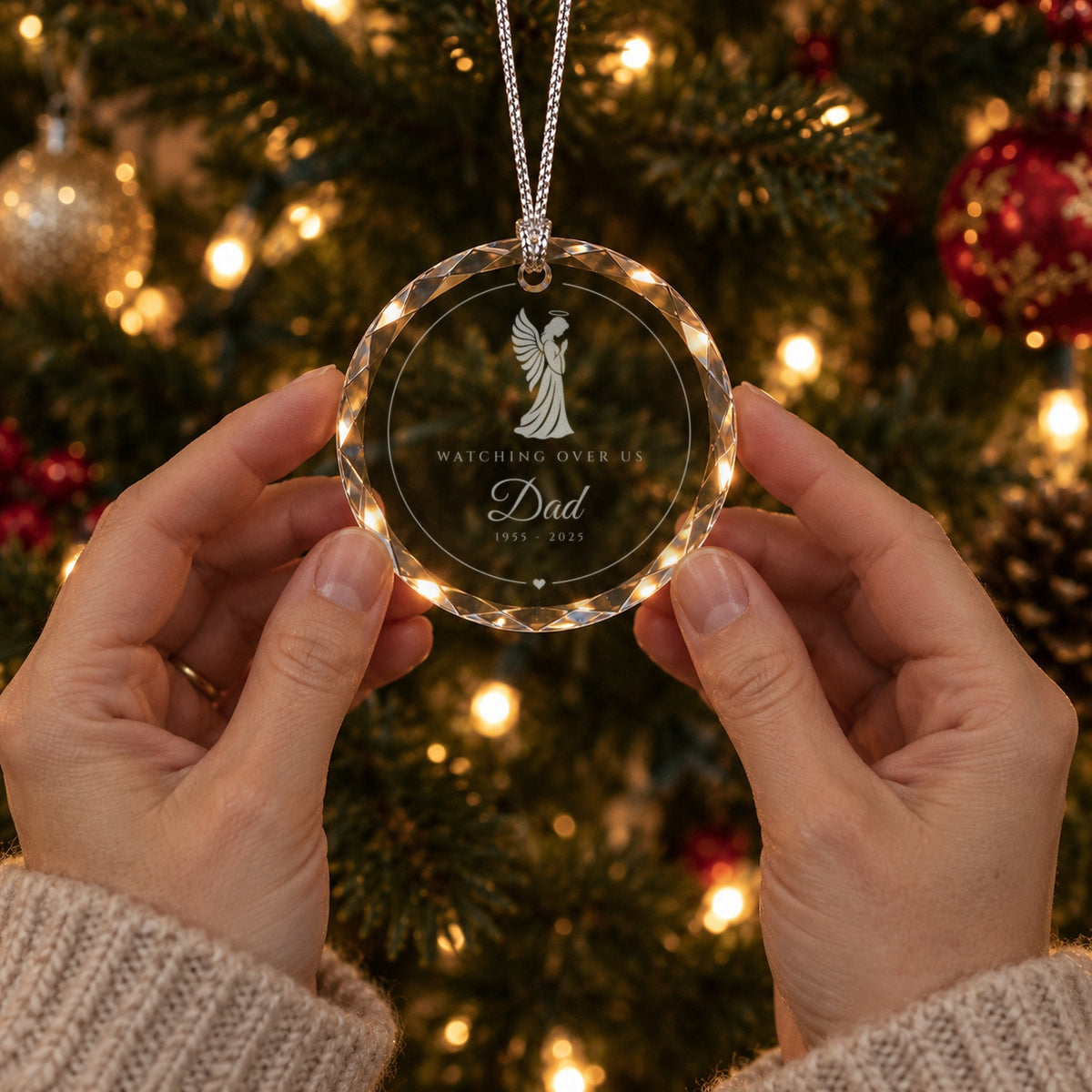 Two hands gently holding the engraved angel memorial ornament in front of a lit Christmas tree, reading Watching Over Us and Dad