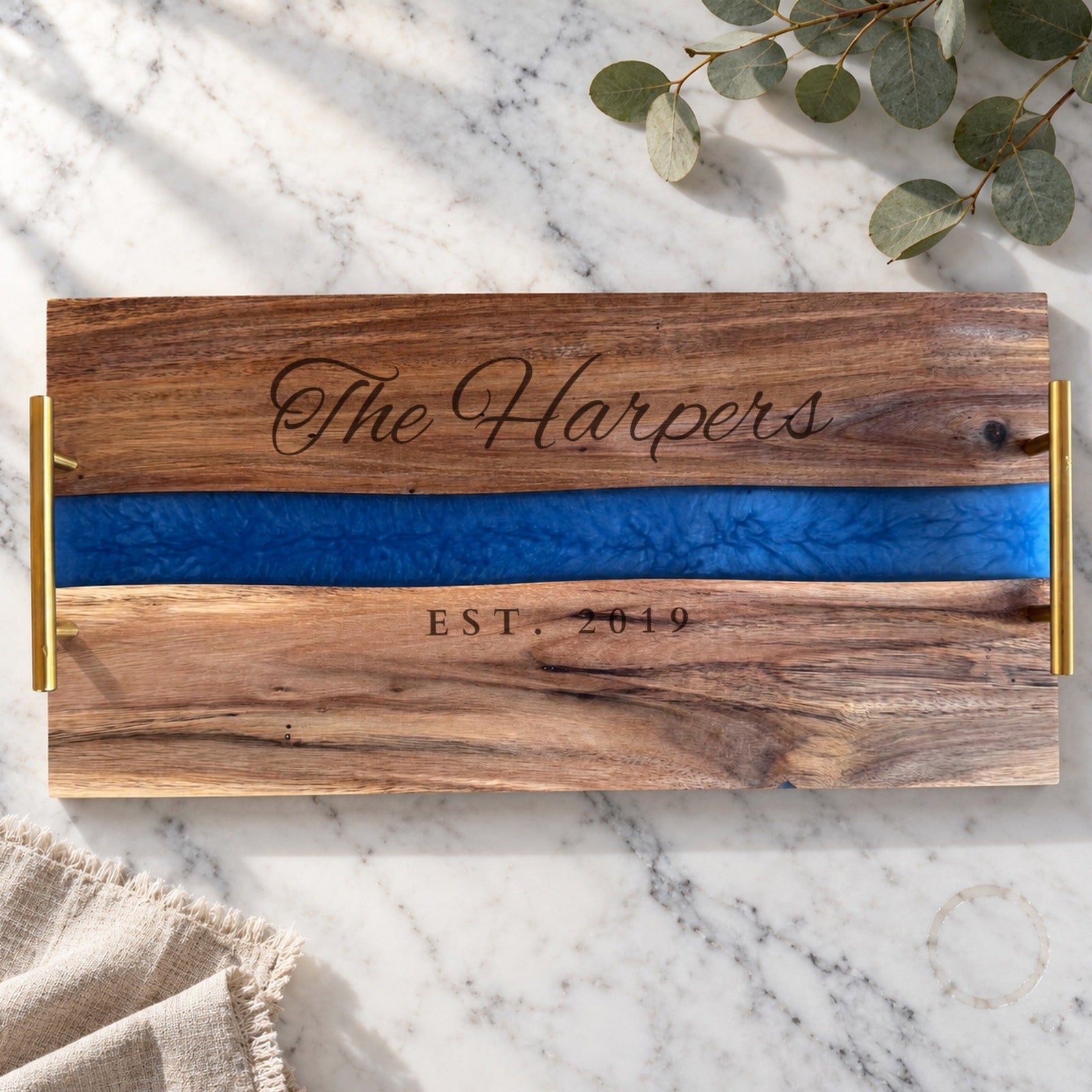 Blue River personalized serving tray engraved The Harpers above a wavy blue resin river and EST. 2019 below it, acacia wood with gold handles on a marble counter