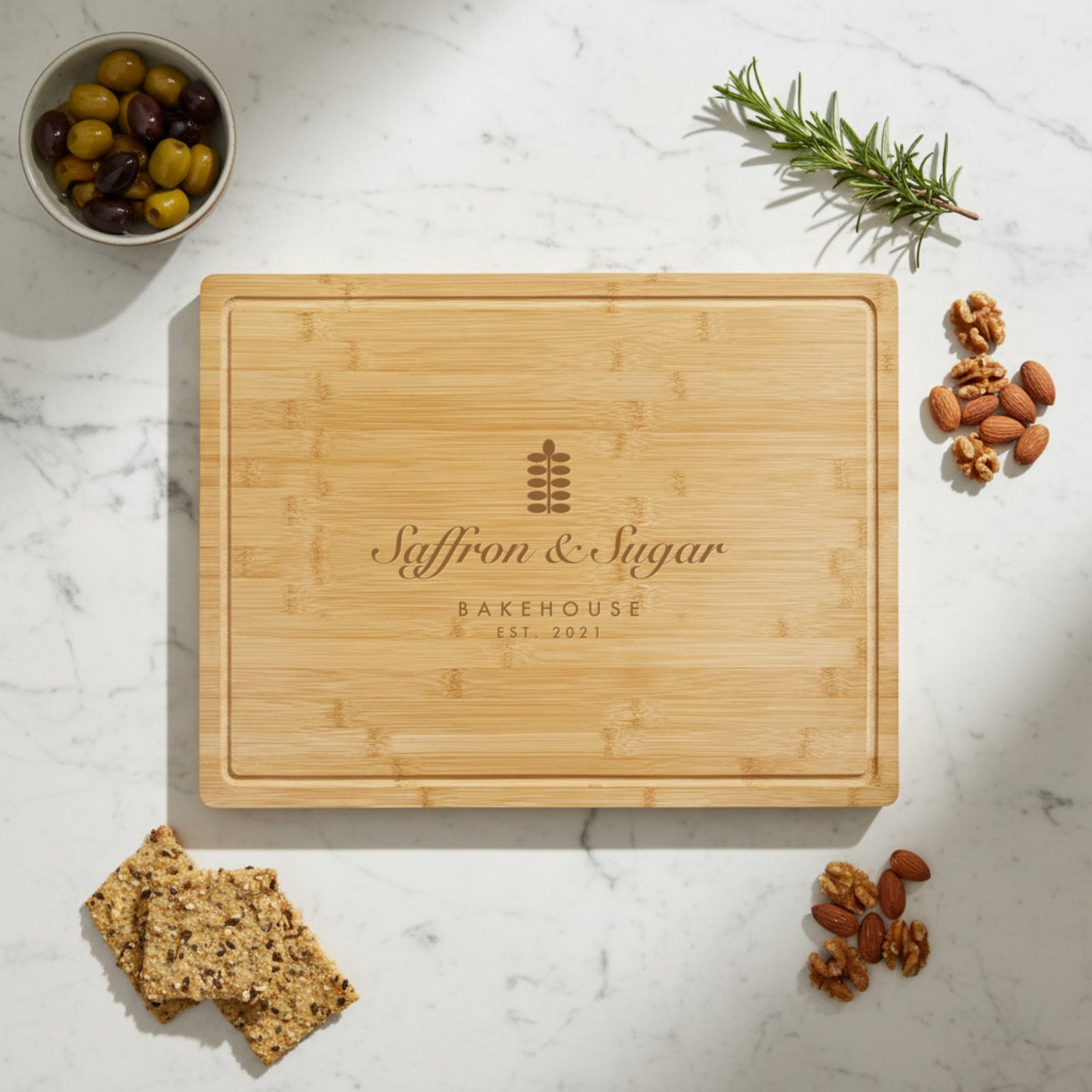 Bamboo cutting board engraved with a sample bakery logo reading Saffron and Sugar Bakehouse, styled on white marble with olives, rosemary and almonds