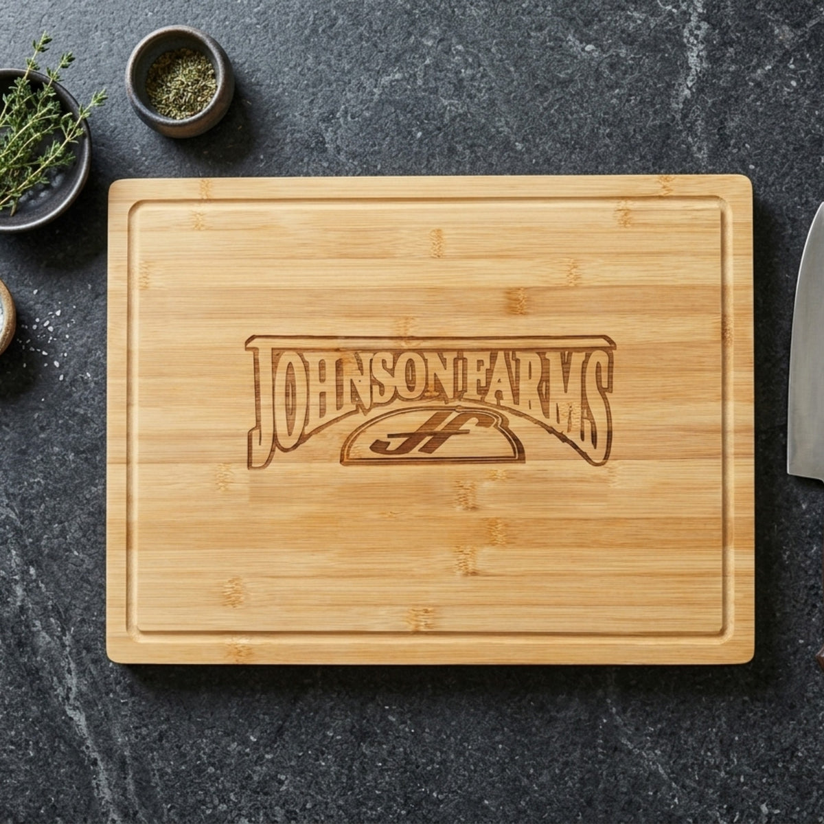 Personalized bamboo cutting board laser engraved with a sample farm business logo, shown on a dark slate kitchen counter with herbs and a chef's knife