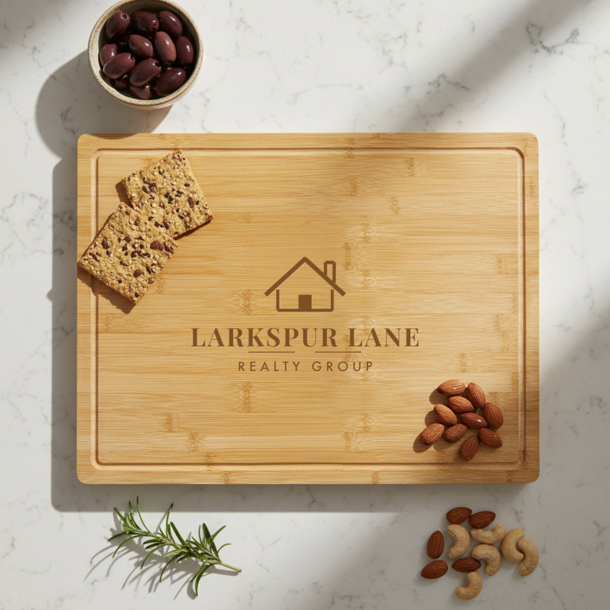 Custom logo cutting board laser engraved with a sample real estate logo, a closing gift for clients shown on a marble counter with olives, crackers and almonds