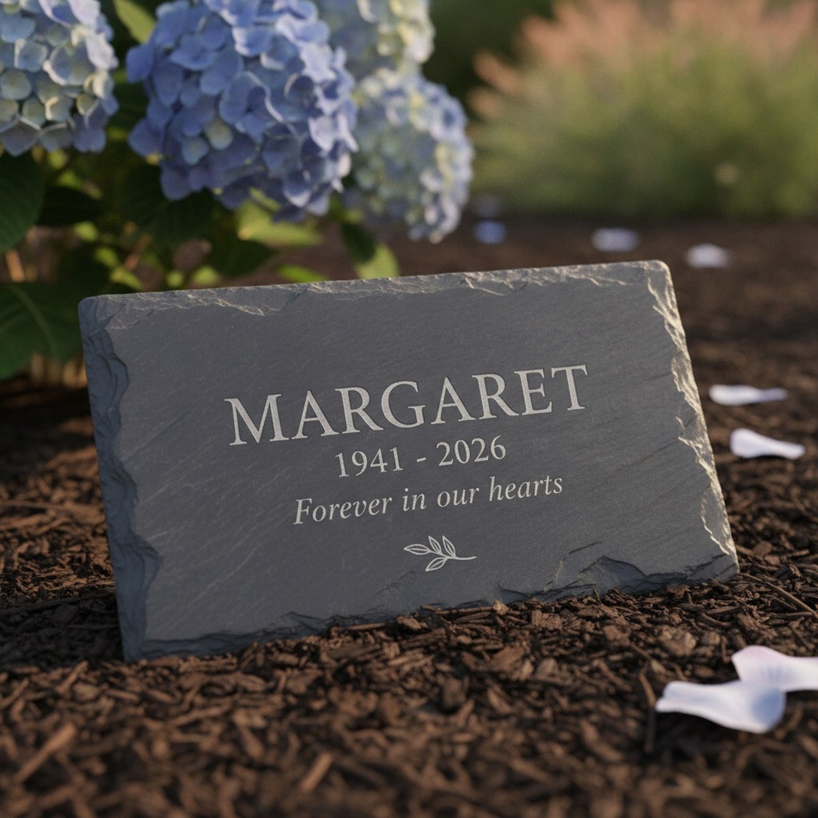 Custom slate memorial garden stone lying on a mulched bed under a blue hydrangea, personalized with a name, dates and the words forever in our hearts
