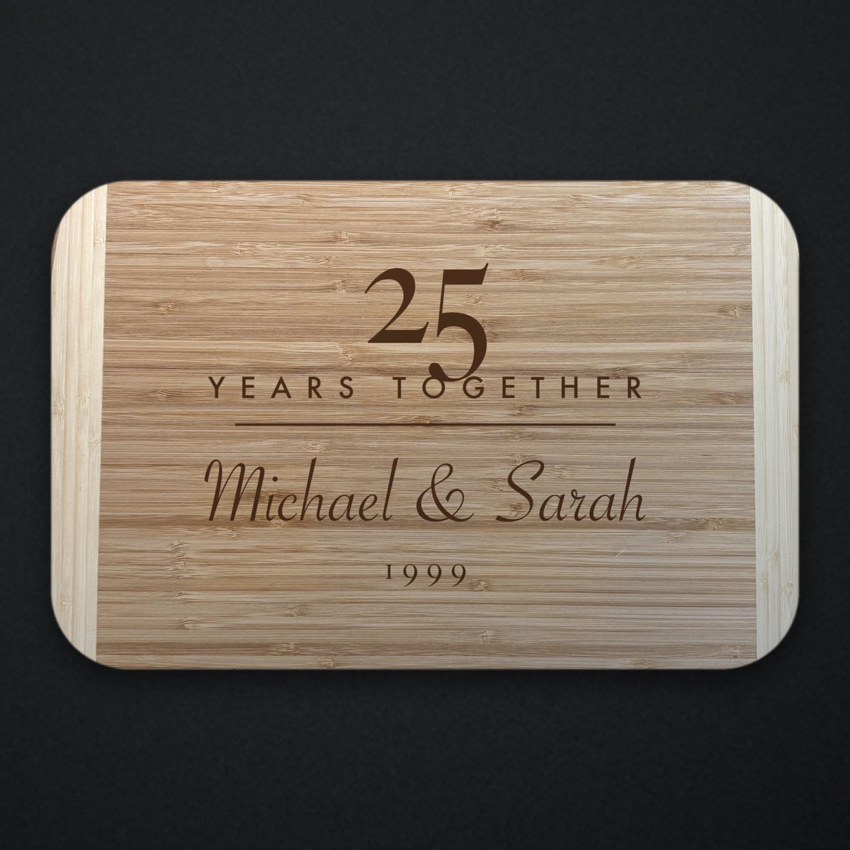 Two tone bamboo anniversary cutting board engraved with a large 25 over YEARS TOGETHER, with Michael and Sarah in script beneath, shown on a dark background.