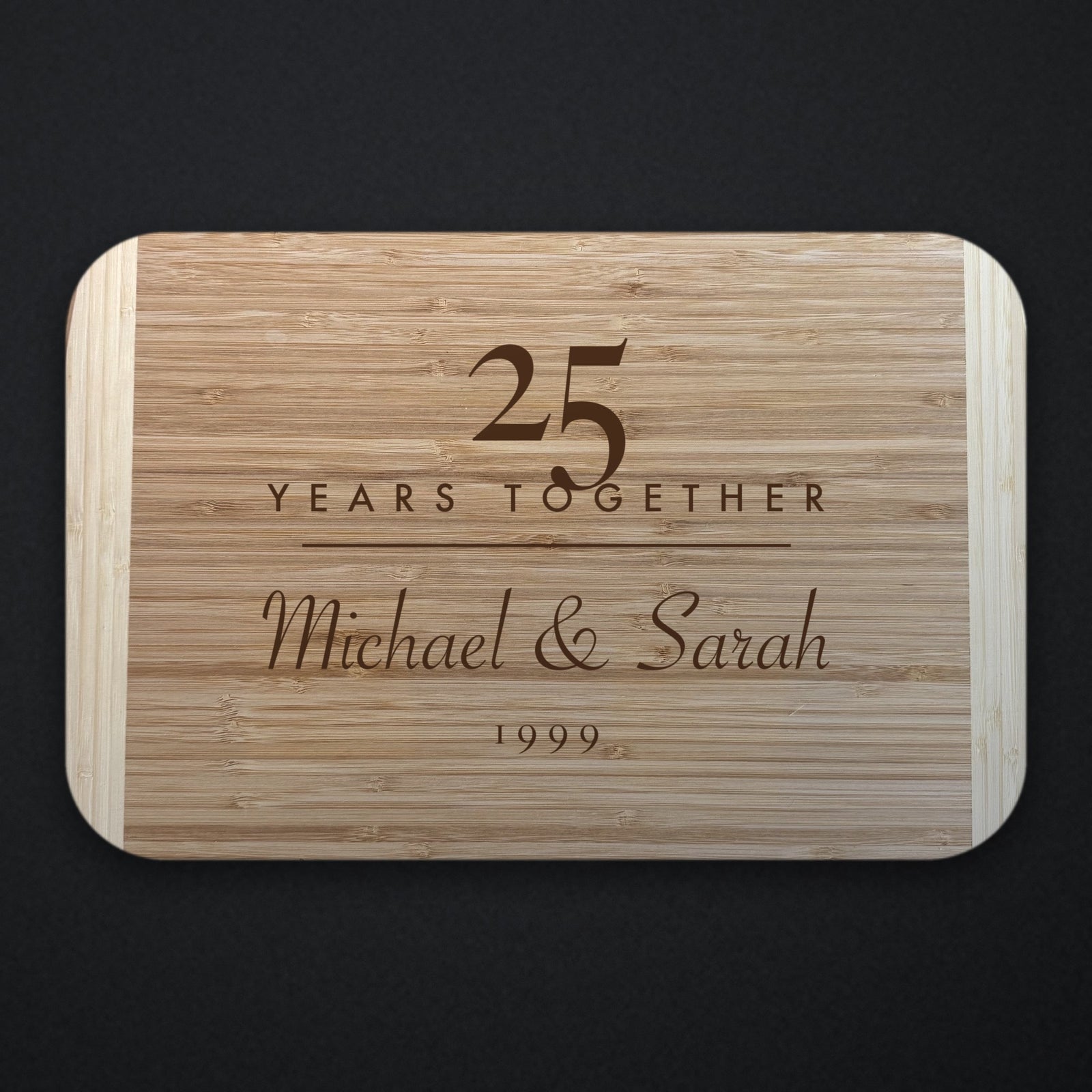 Two tone bamboo anniversary cutting board engraved with a large 25 over YEARS TOGETHER, with Michael and Sarah in script beneath, shown on a dark background.