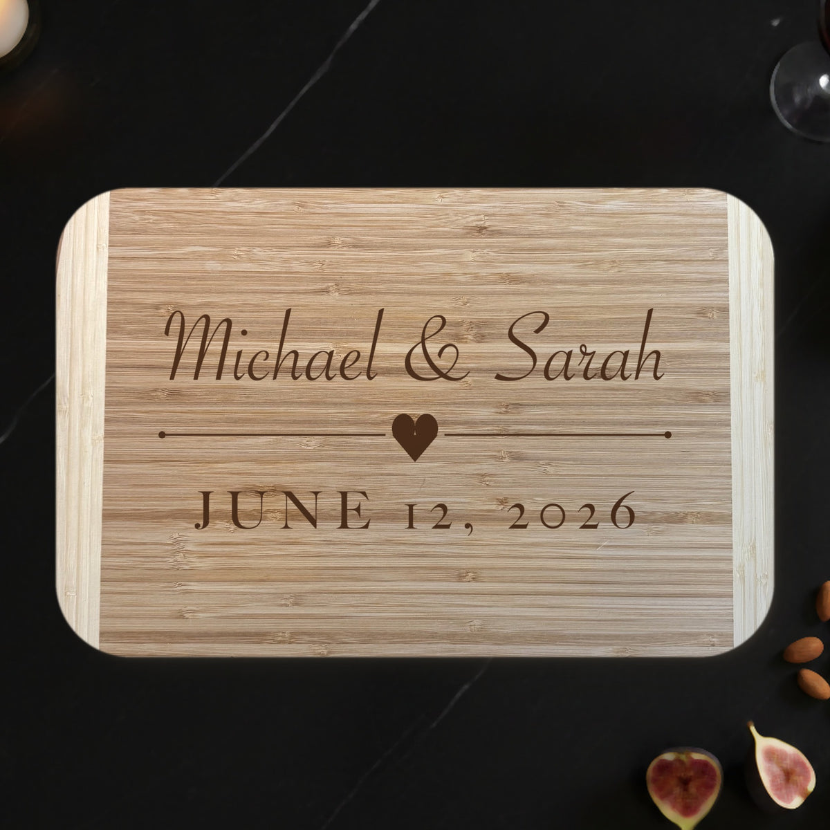 Personalized couples cutting board on a black marble countertop with wine, figs and a candle, engraved Michael and Sarah above a heart with JUNE 12 2026 on two tone bamboo.