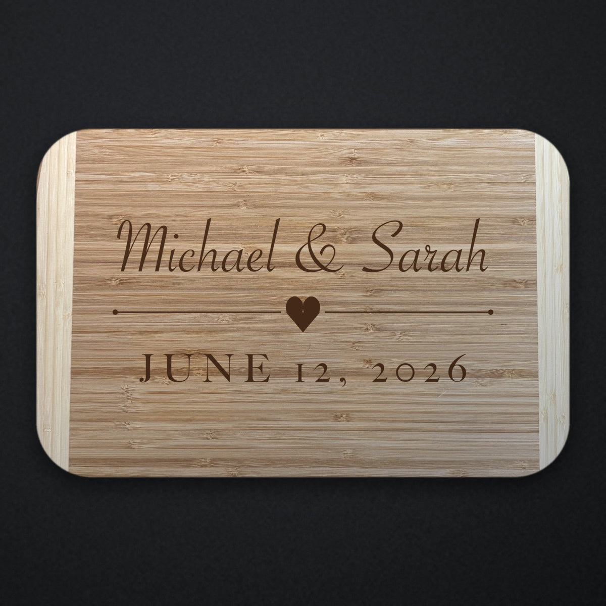 Two tone bamboo couples cutting board engraved with Michael and Sarah in script above a heart and rule, with JUNE 12 2026 beneath, shown on a dark background.