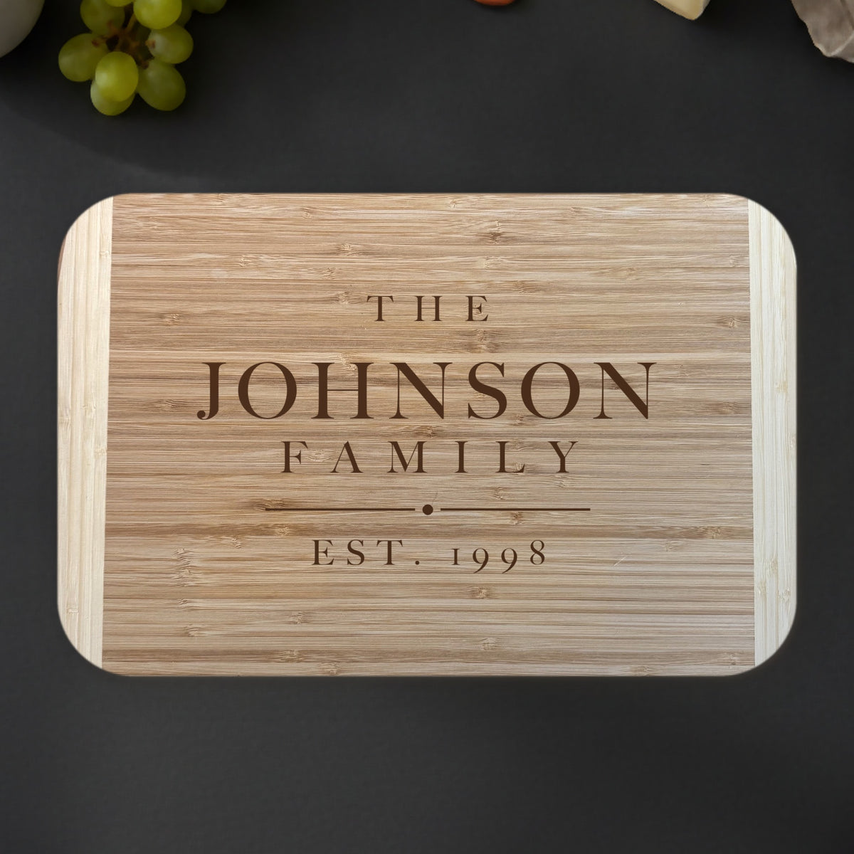 Personalized family name cutting board on a dark slate kitchen counter with grapes and eucalyptus, engraved THE JOHNSON FAMILY EST. 1998 on two tone bamboo.