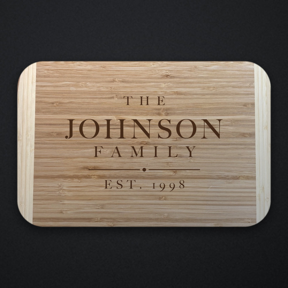 Two tone bamboo cutting board engraved THE JOHNSON FAMILY with EST. 1998 beneath a rule and centre dot, shown on a dark background.