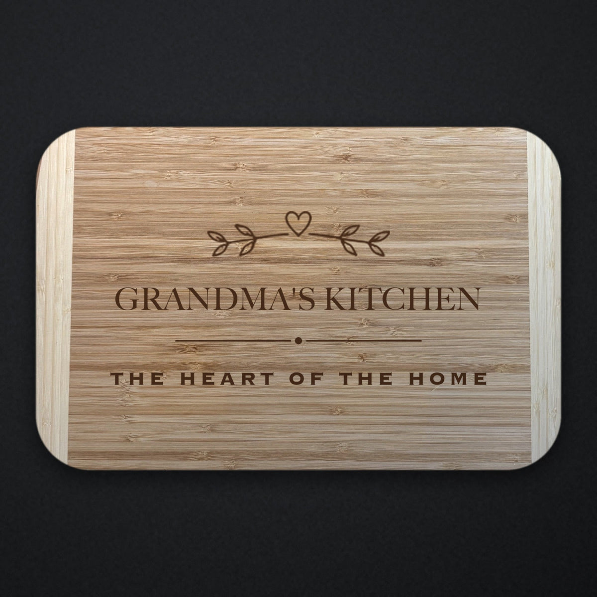 Two tone bamboo cutting board engraved GRANDMA'S KITCHEN under a sprig and heart, with THE HEART OF THE HOME on the line below a rule, on a dark background.