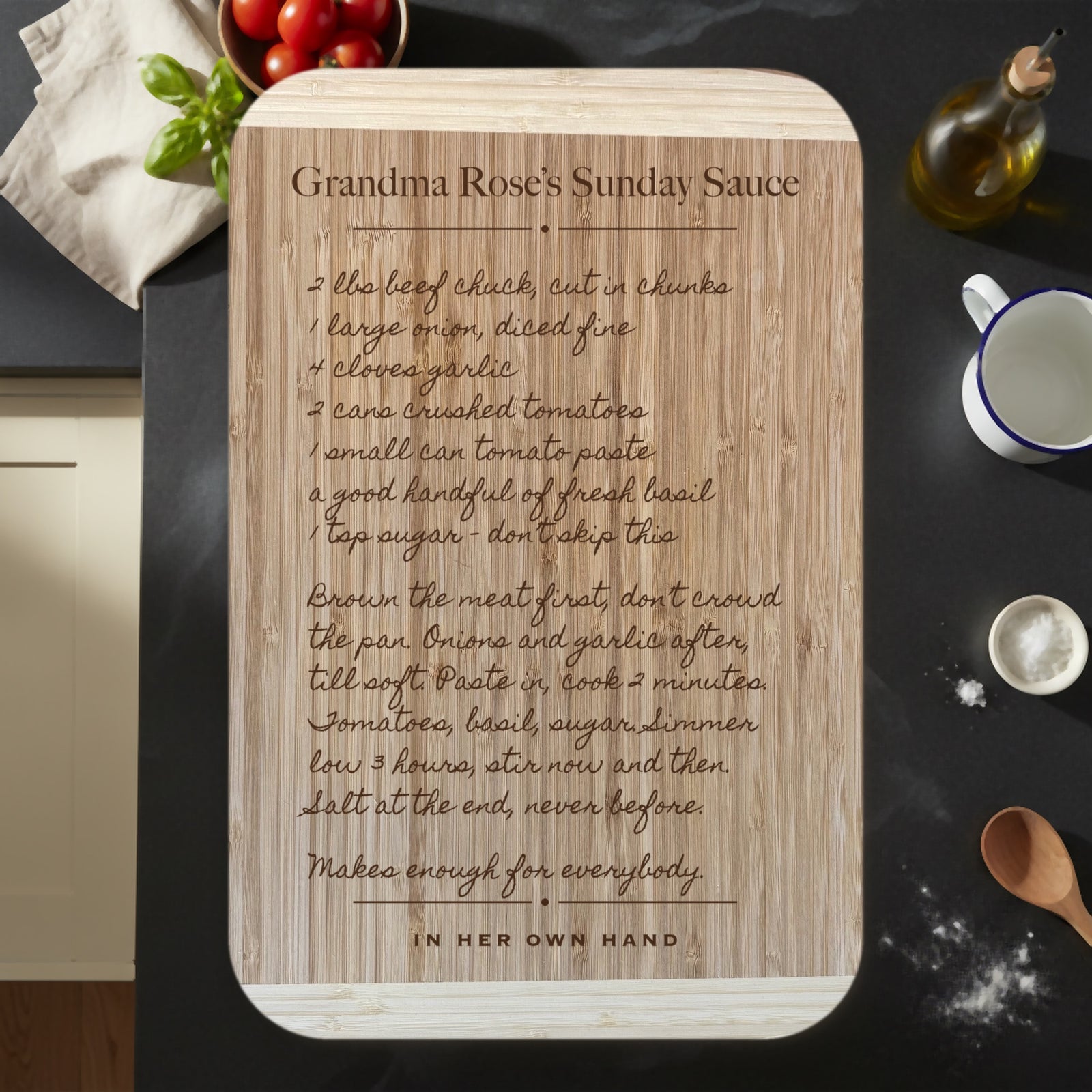 Personalized handwritten recipe cutting board on a dark kitchen counter, a family sauce recipe engraved in cursive on two tone bamboo beside tomatoes, basil and olive oil.