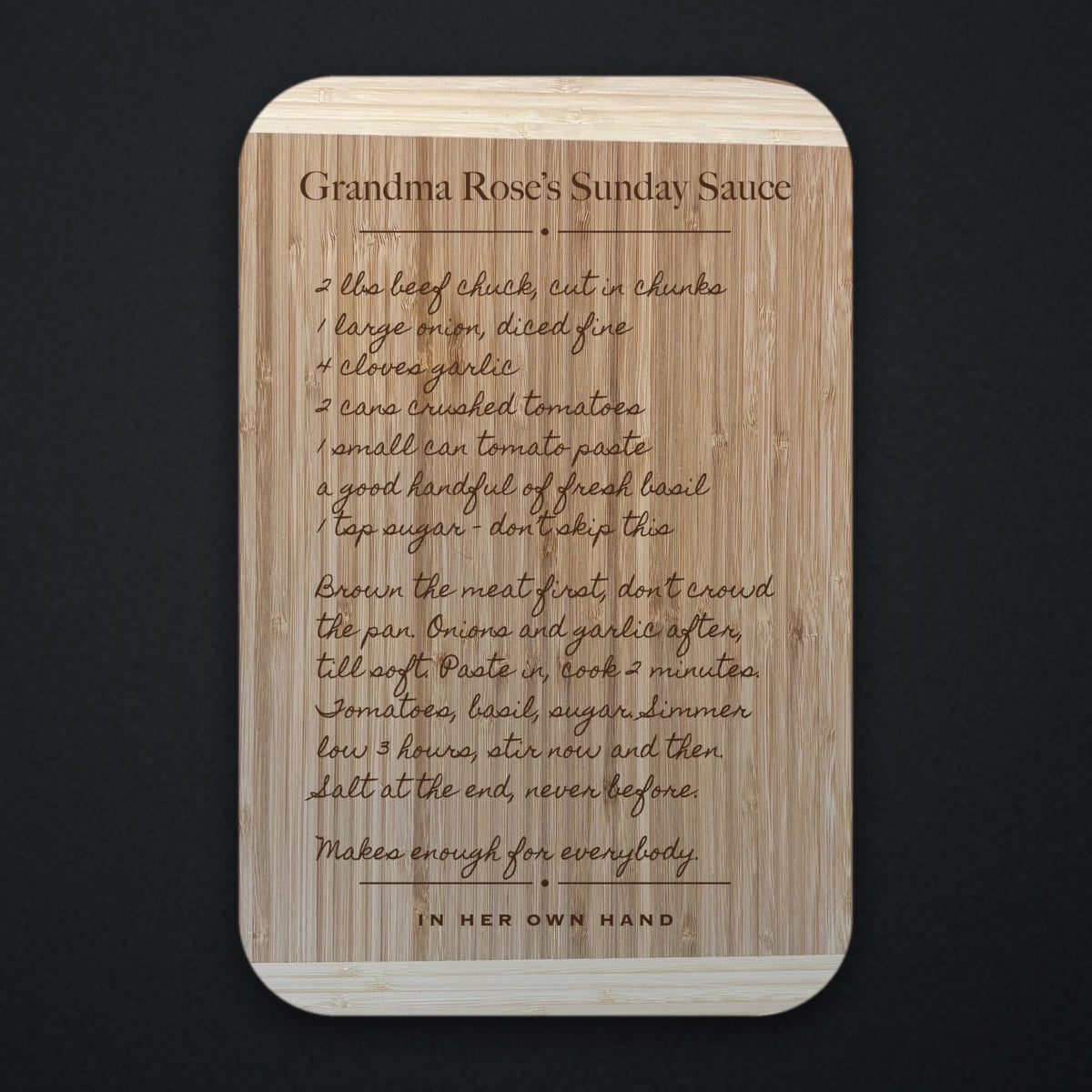 Two tone bamboo recipe cutting board engraved with a handwritten family recipe and the title Grandma Rose’s Sunday Sauce, photographed straight on against a dark background.