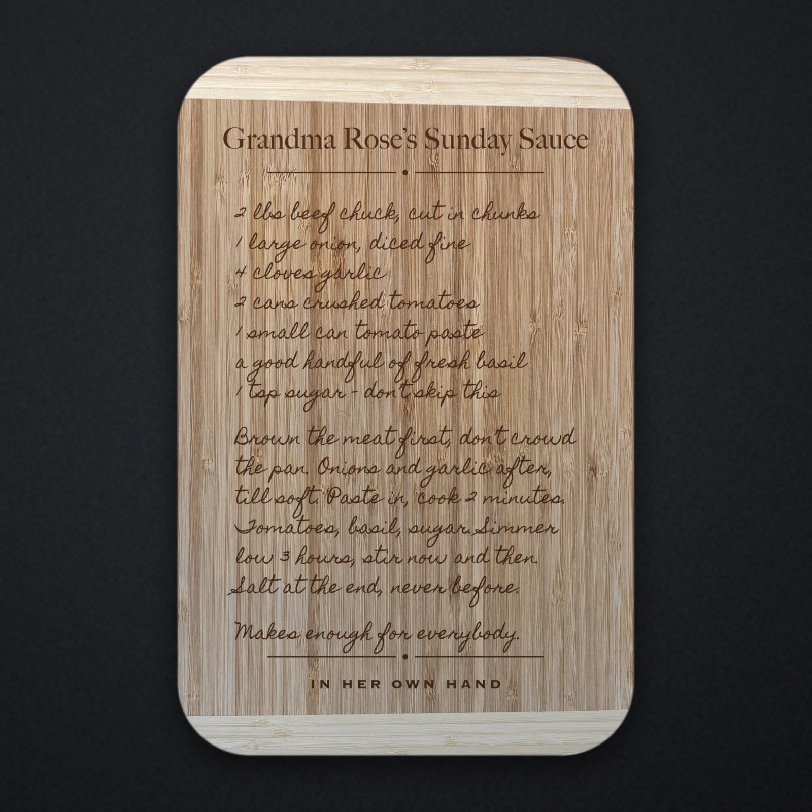Two tone bamboo recipe cutting board engraved with a handwritten family recipe and the title Grandma Rose’s Sunday Sauce, photographed straight on against a dark background.