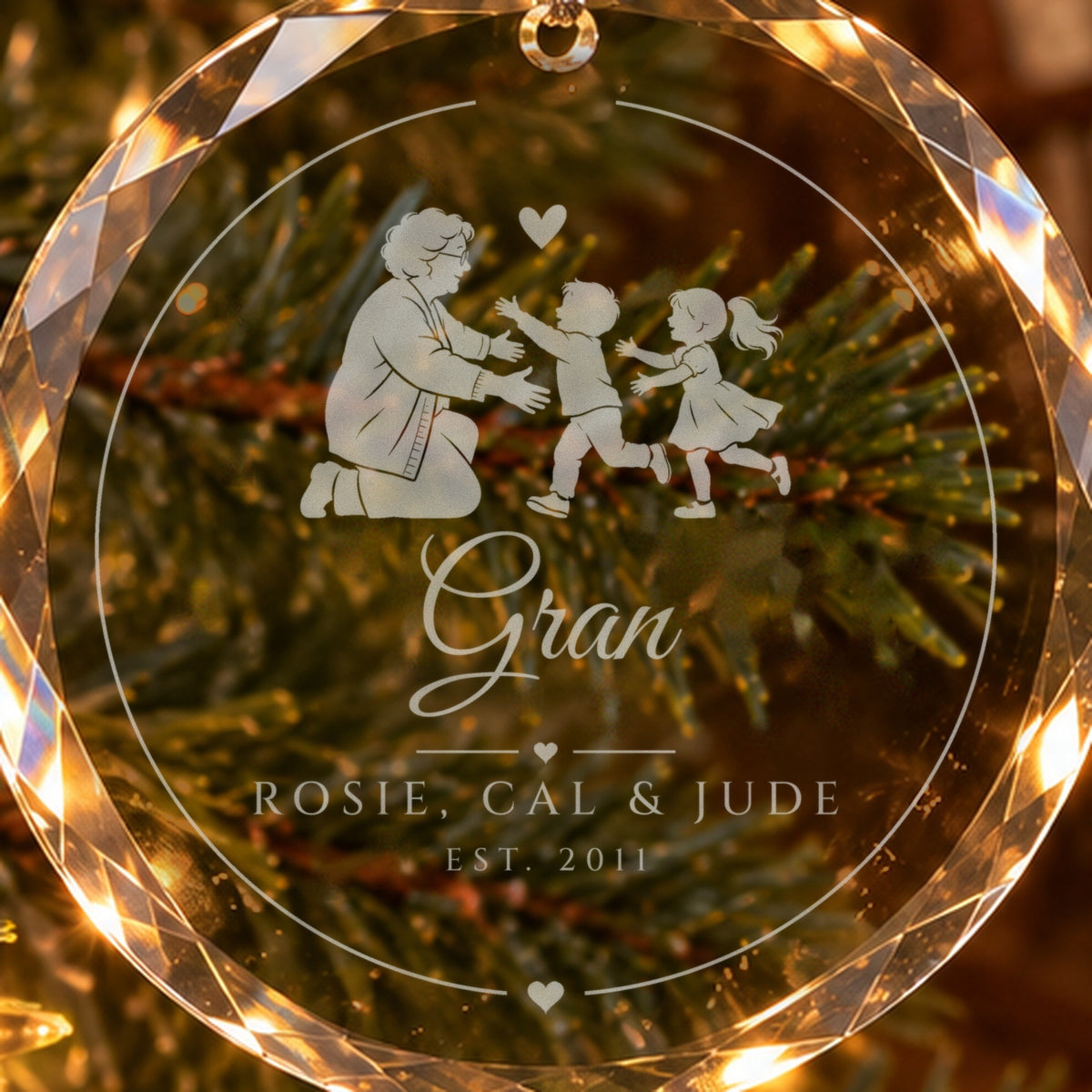 Close-up of the frosted engraving on the grandma ornament: a grandmother opening her arms to two running grandkids, Gran in script, ROSIE, CAL & JUDE, EST. 2011