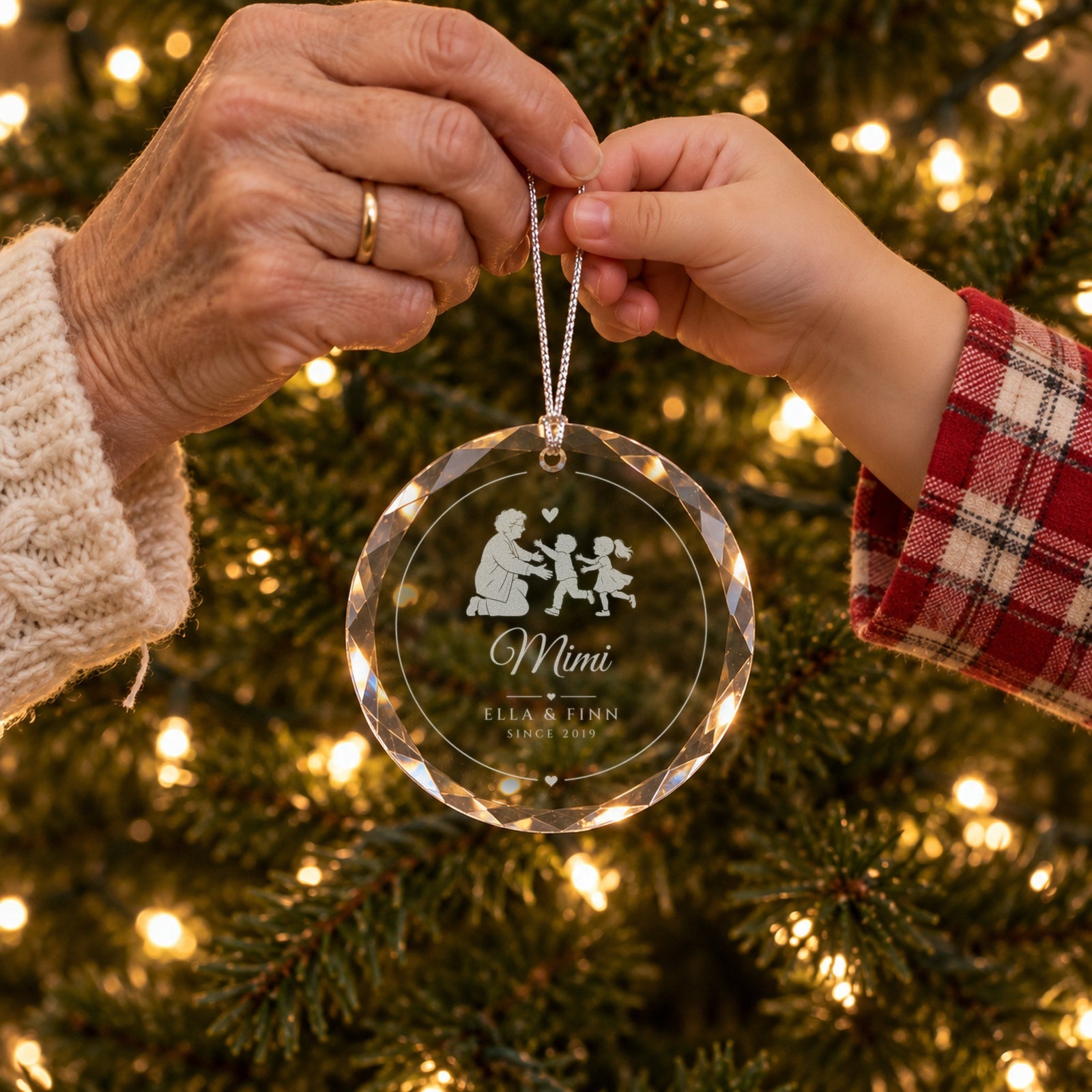 A grandmother's hand and a grandchild's hand holding the engraved glass ornament together by the tree, Mimi in script with ELLA & FINN, SINCE 2019
