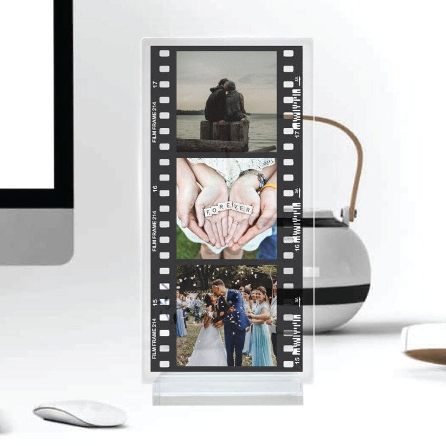 Clear acrylic photo display shaped like a film strip showing three sample photos, standing on a desk near a monitor