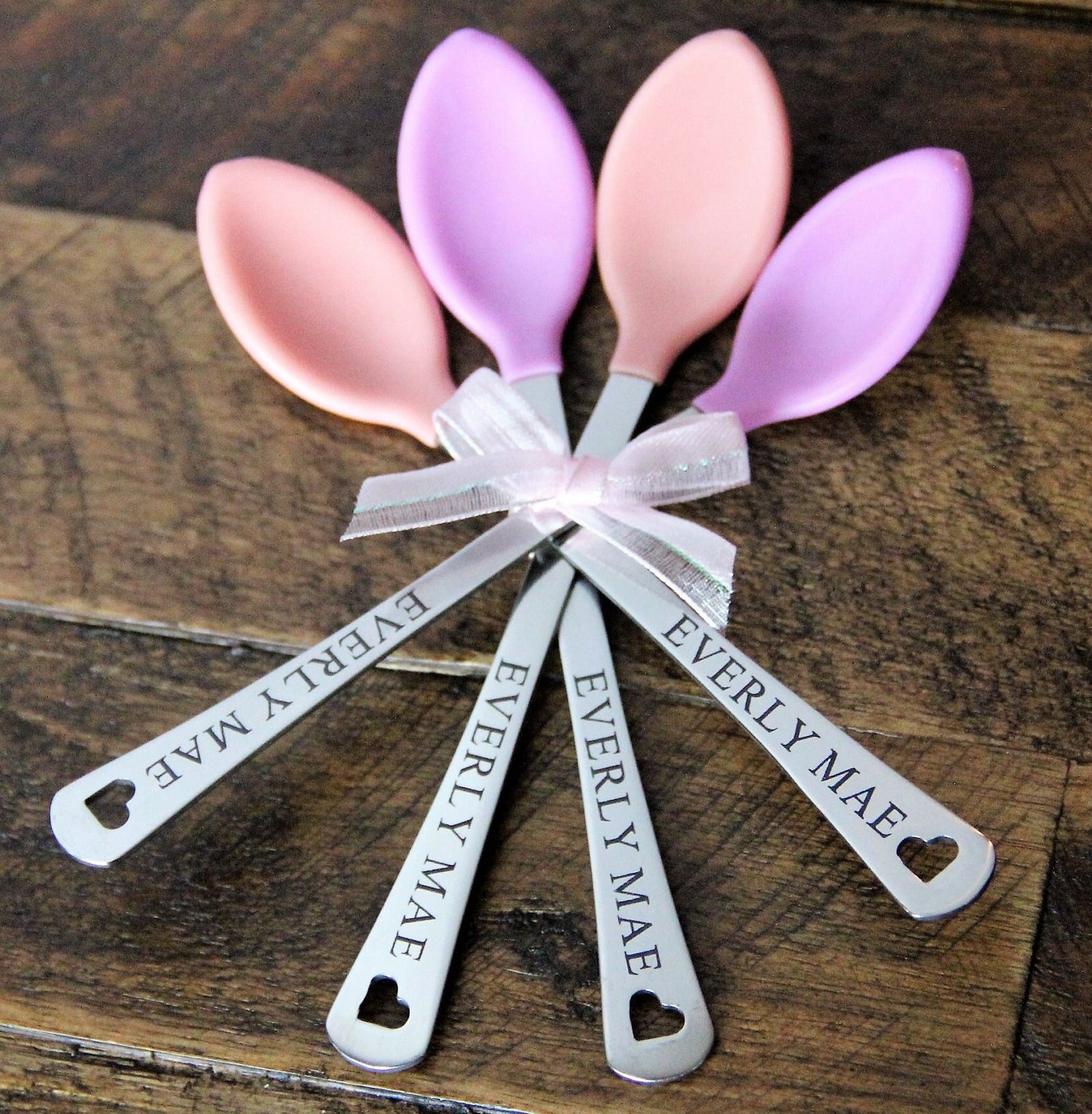 Engraved baby girl spoon set hero photo showing four pink and peach silicone baby spoons with personalized name engravings, styled on a soft neutral background for a baby shower gift.