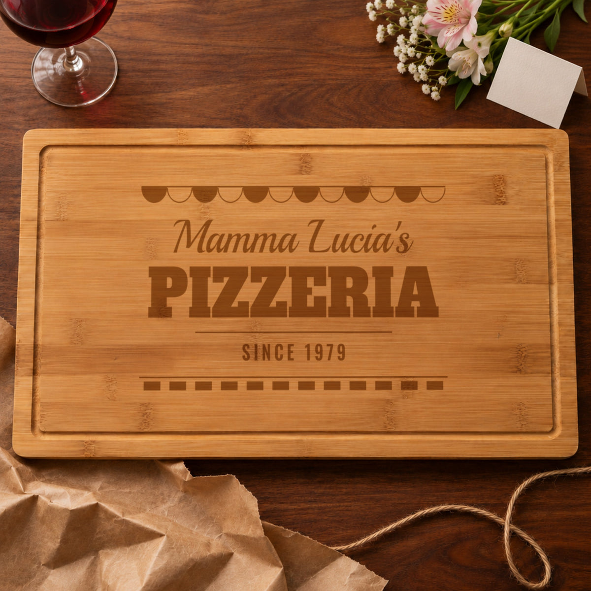 Personalized Italian pizzeria board engraved Mamma Lucia's PIZZERIA SINCE 1979, unwrapped from kraft paper beside flowers and red wine, a gift for grandma