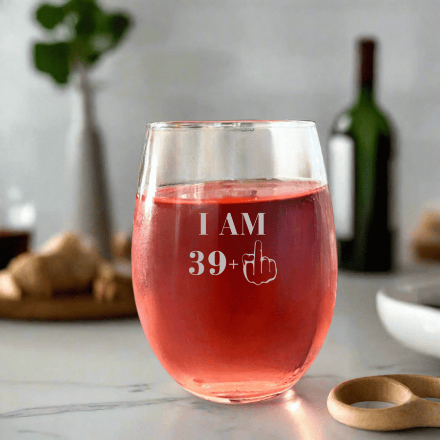 40th birthday stemless wine glass engraved 'I am 39+' with a middle finger graphic, filled with rosé wine indoors