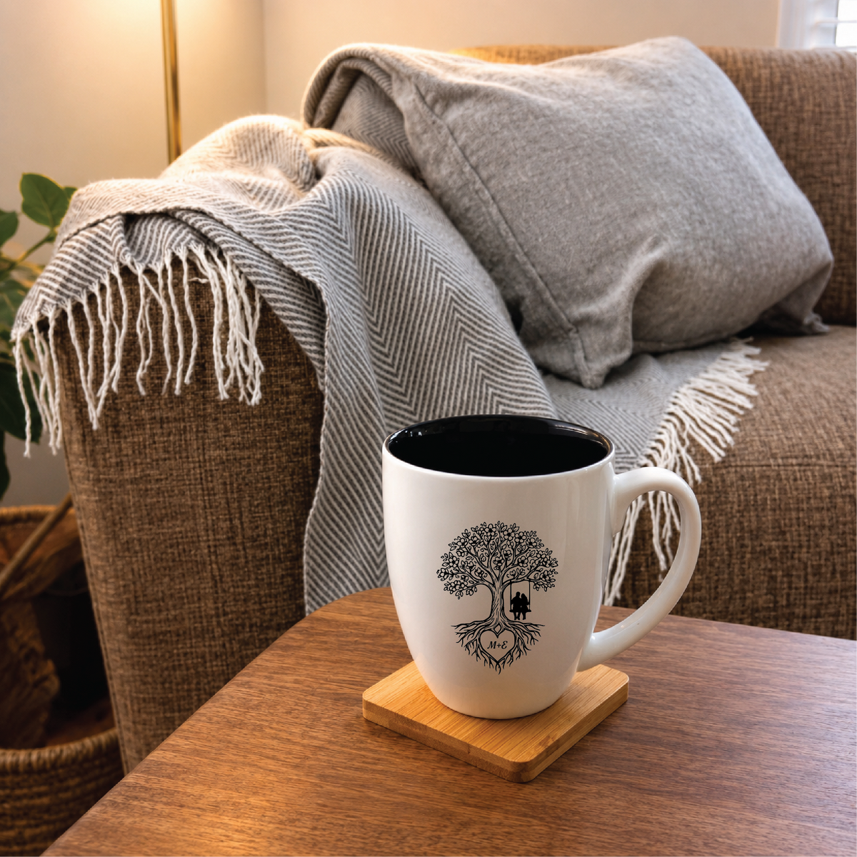 Engraved couples mug with a tree of life and two initials resting on a bamboo coaster beside a grey fringed throw blanket on an armchair.