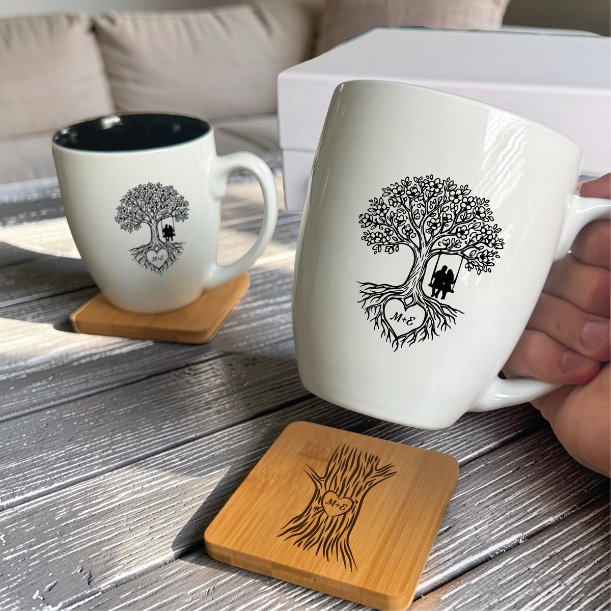 Two engraved bistro mugs and two bamboo coasters, the mugs carrying a tree of life with a couple on a swing and the coasters a carved trunk holding the couple initials.
