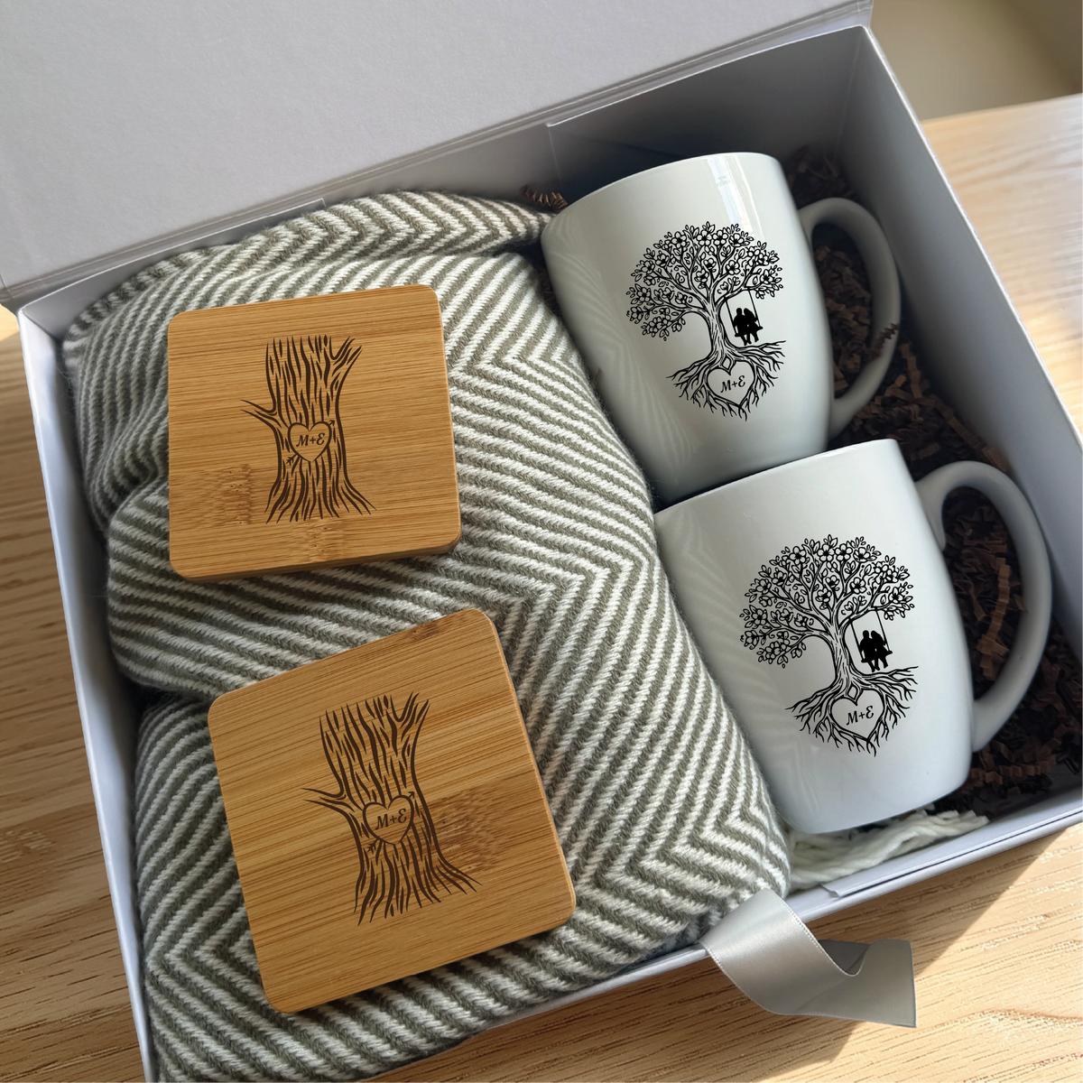 Personalized couples gift set open in a keepsake box with two engraved tree of life mugs, two bamboo coasters carved with their initials and a grey herringbone blanket.