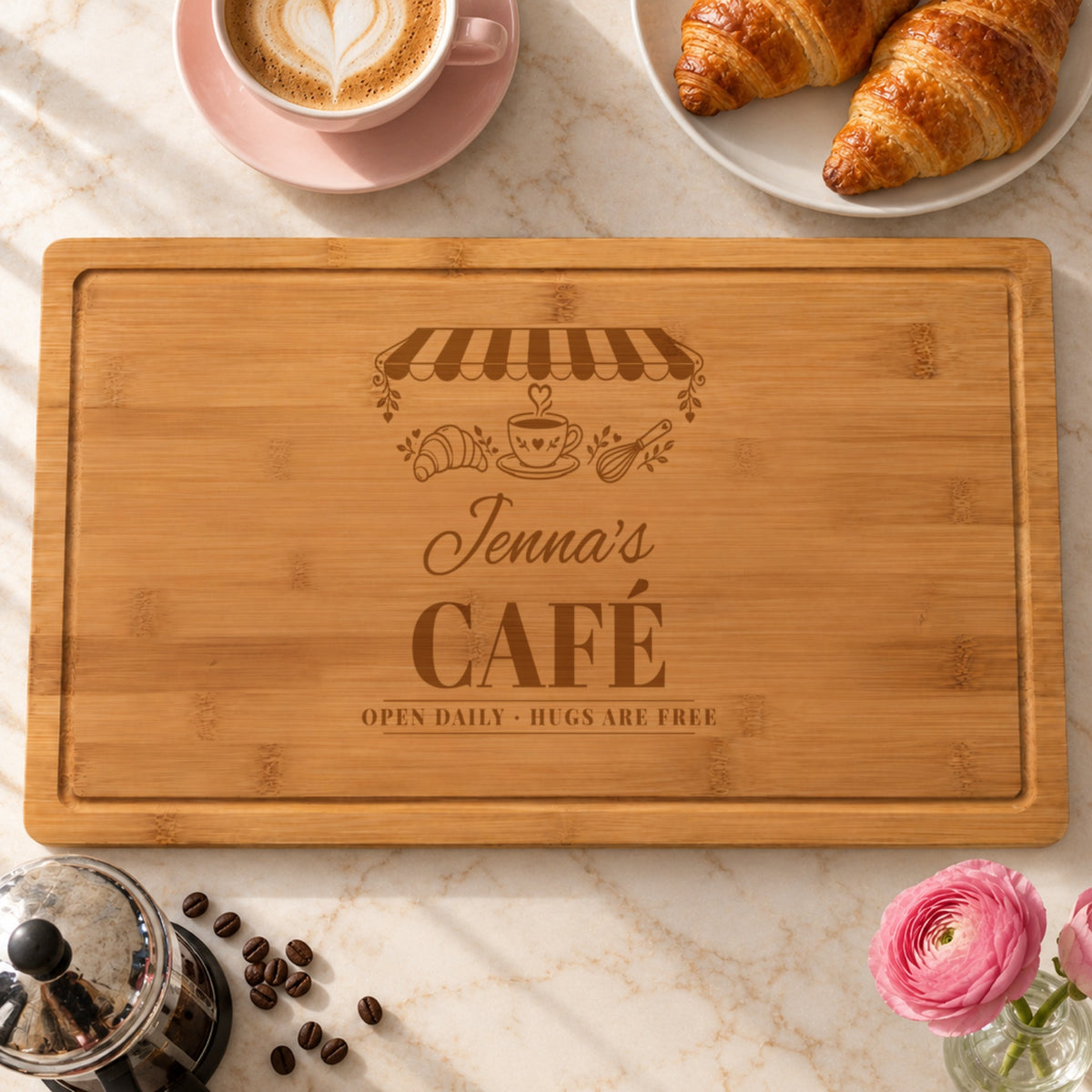Coffee lover cutting board engraved like a vintage cafe sign, Jenna's CAFÉ OPEN DAILY HUGS ARE FREE, with a striped awning and heart coffee cup, beside a latte and croissants