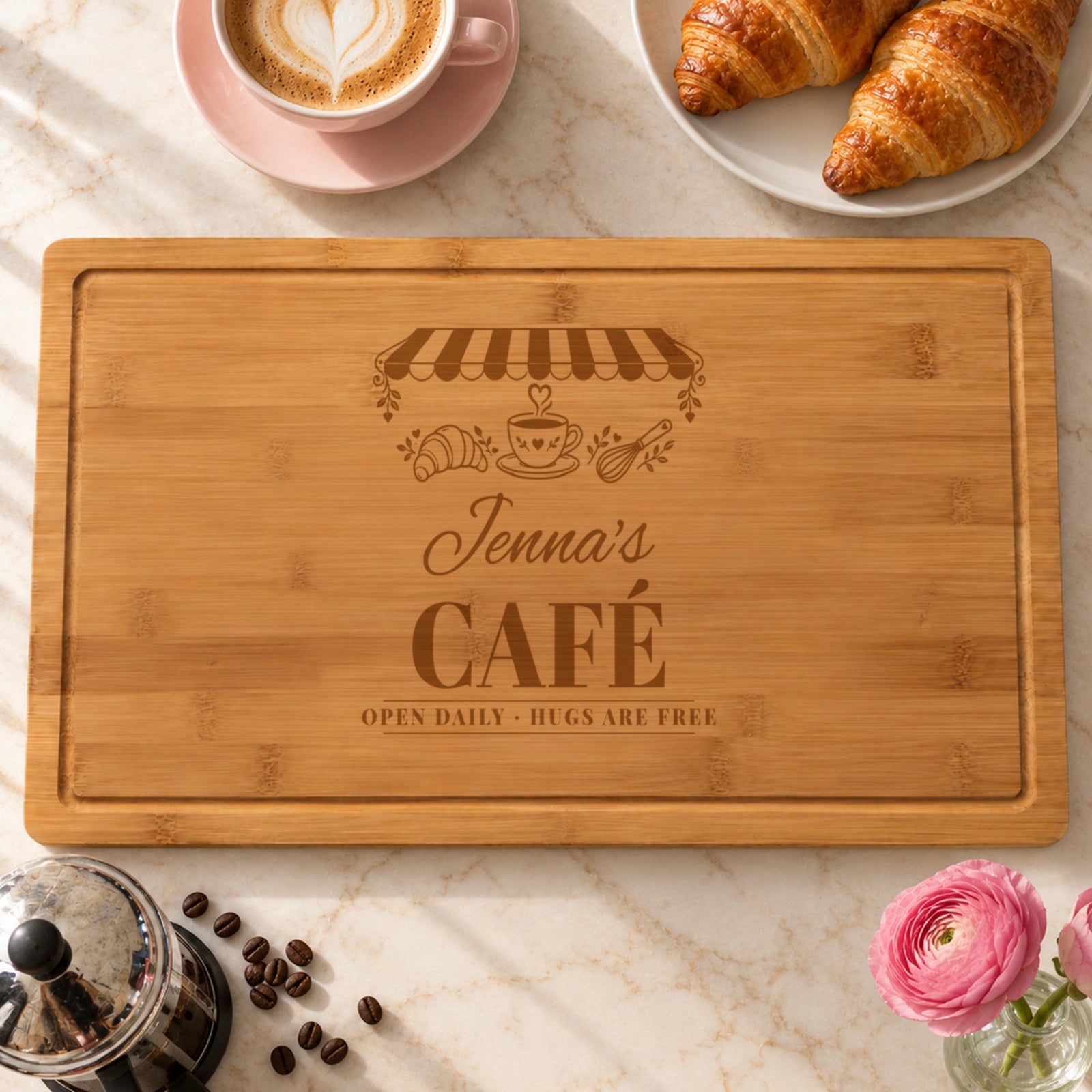 Coffee lover cutting board engraved like a vintage cafe sign, Jenna's CAFÉ OPEN DAILY HUGS ARE FREE, with a striped awning and heart coffee cup, beside a latte and croissants