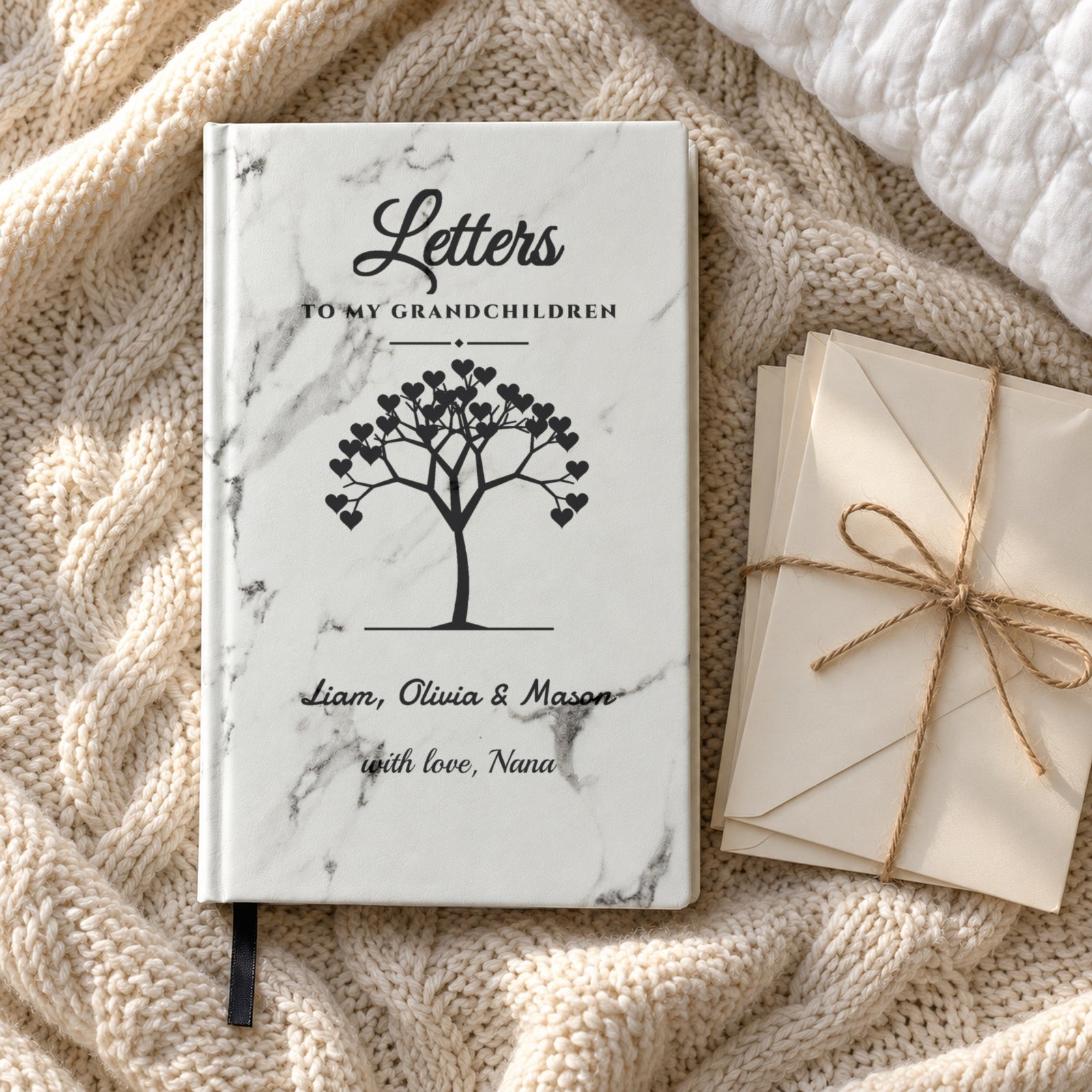 Personalized grandma journal engraved Liam, Olivia & Mason and with love, Nana, on a knit blanket beside blank envelopes tied with twine, a Christmas gift for grandma
