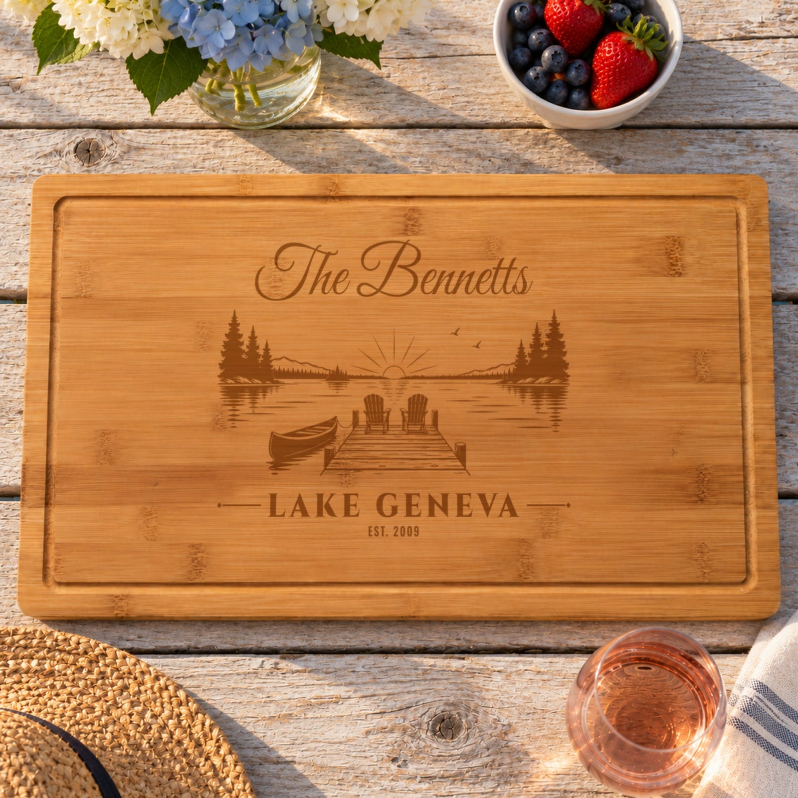 Personalized lake house sign cutting board engraved The Bennetts, Lake Geneva, EST. 2009 over a sunset dock, with hydrangeas, berries and rosé on a cottage porch table