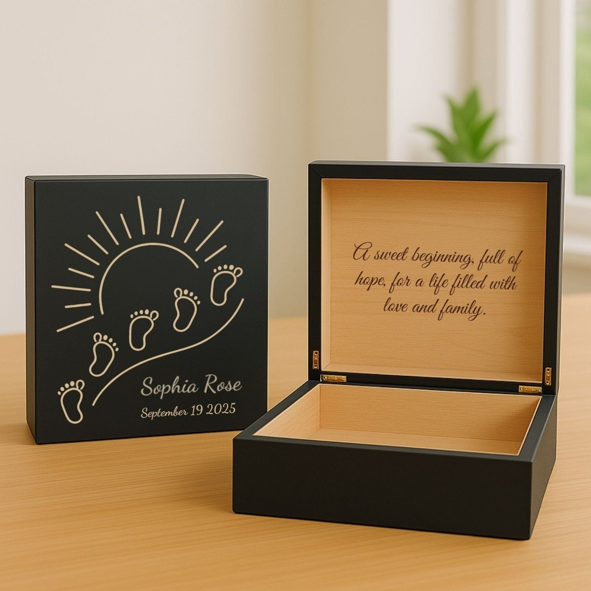 Black personalized newborn keepsake box with light engraved baby footprints, a rising sun and a sample name and date, next to the open box and its message
