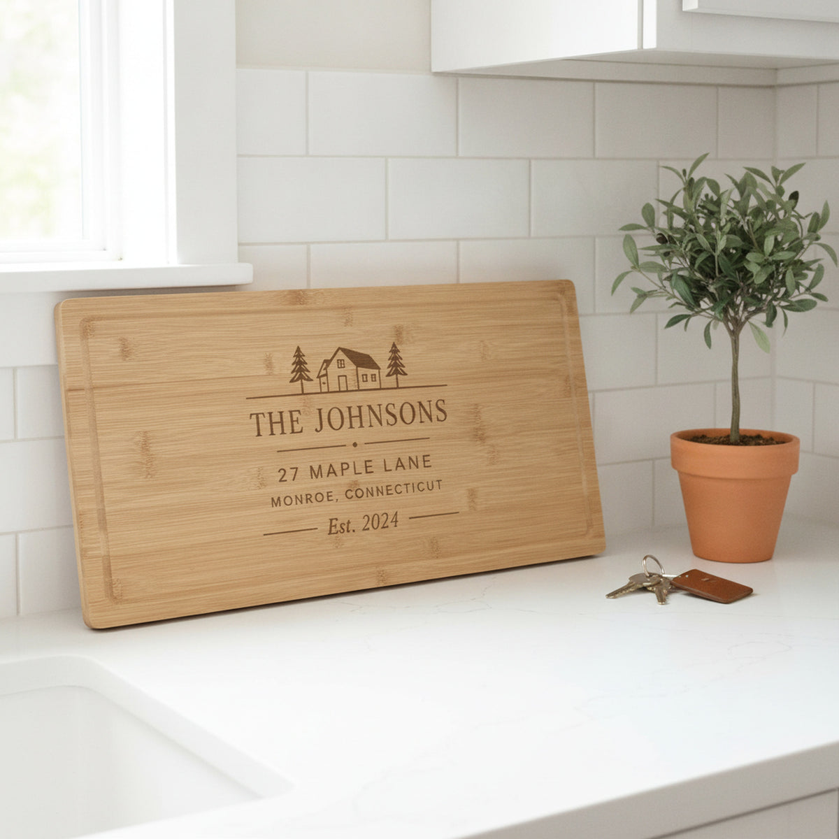 Engraved address board leaning on a white quartz counter in a bright new kitchen beside house keys on a leather fob and a potted olive tree.