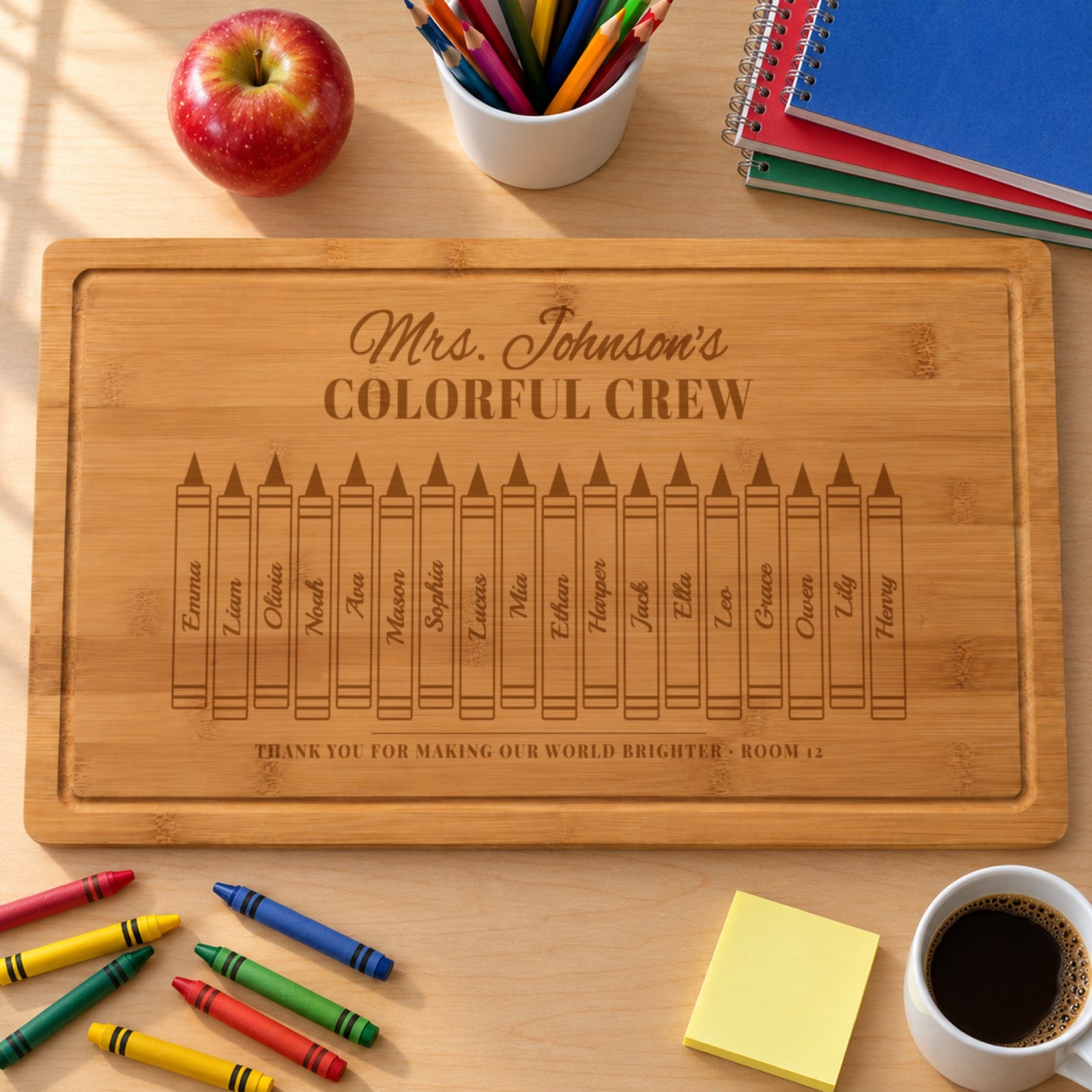 Personalized teacher gift cutting board engraved Mrs. Johnson's COLORFUL CREW with eighteen student names on crayons, on a teacher desk with an apple