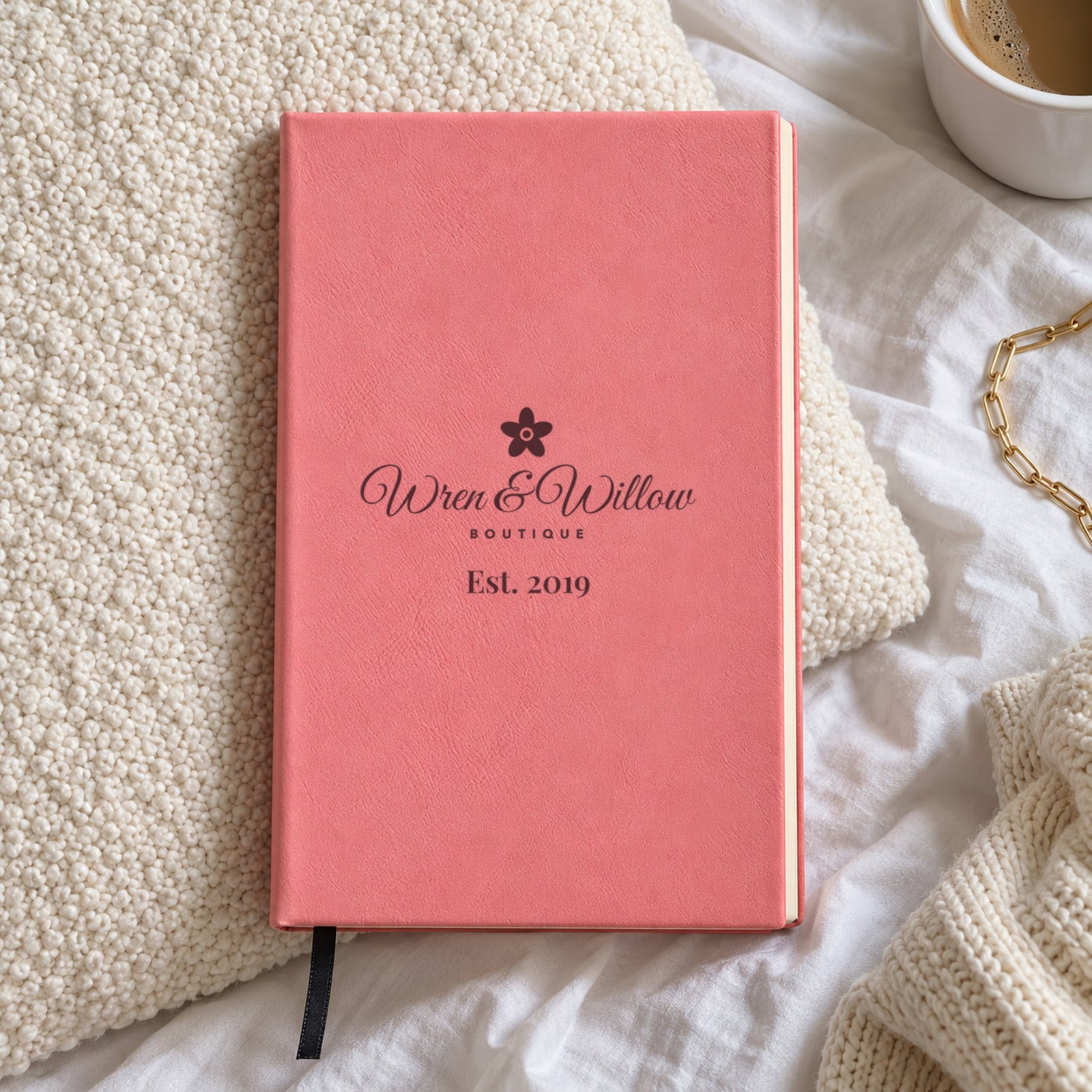 Pink engraved journal with a boutique logo, a flower over Wren & Willow in script and Est. 2019, lying on a cream knit pillow on a bed