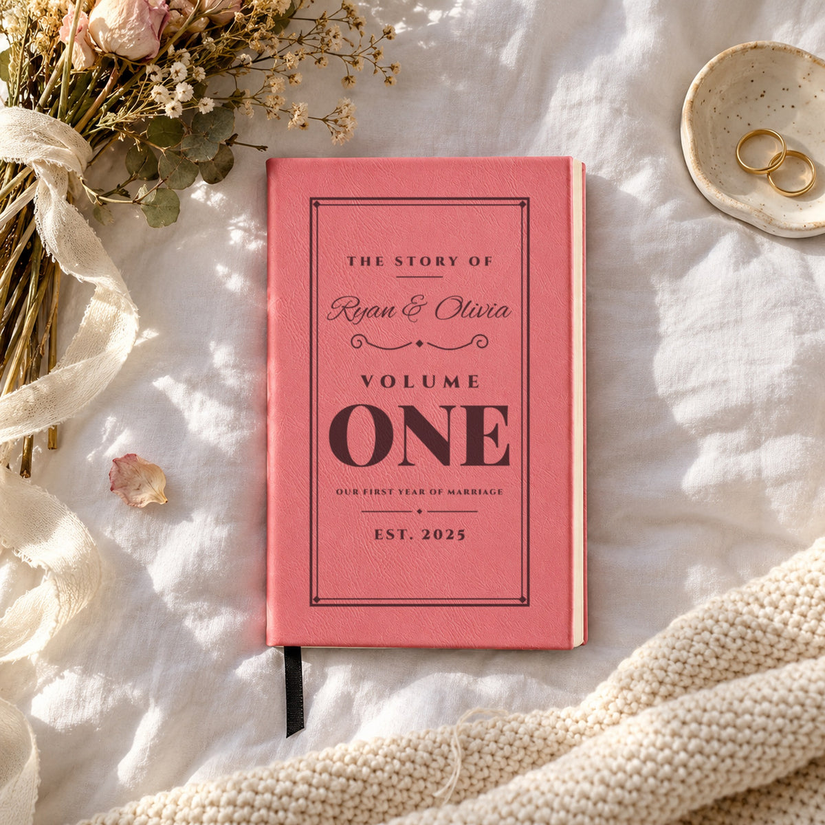 Pink personalized couples journal engraved The Story Of Ryan & Olivia, Volume One, Est. 2025, on a bed beside wedding rings and a dried bouquet