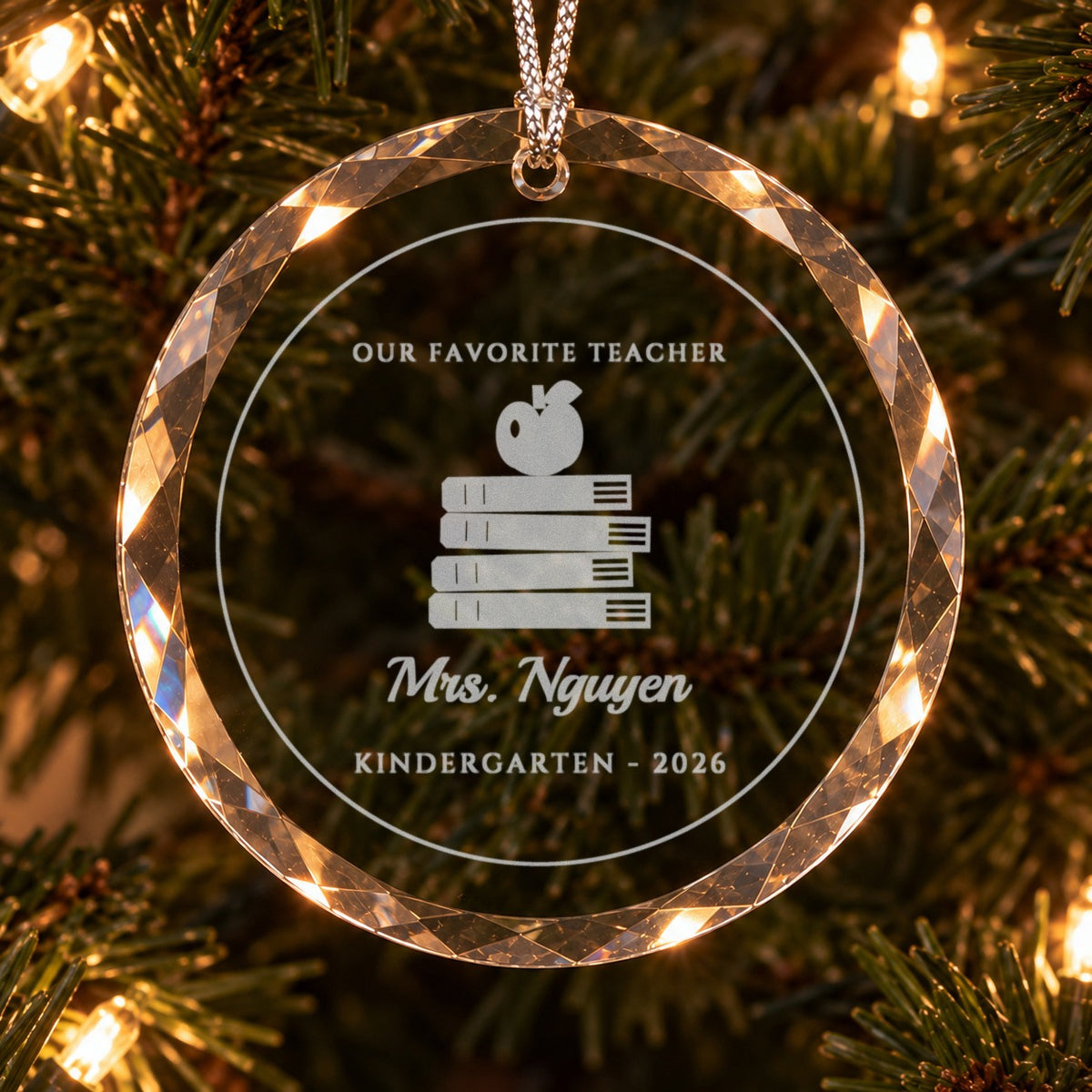 Close-up of the frosted engraving on the teacher ornament: OUR FAVORITE TEACHER, a stack of books with an apple, Mrs. Nguyen in script and KINDERGARTEN 2026
