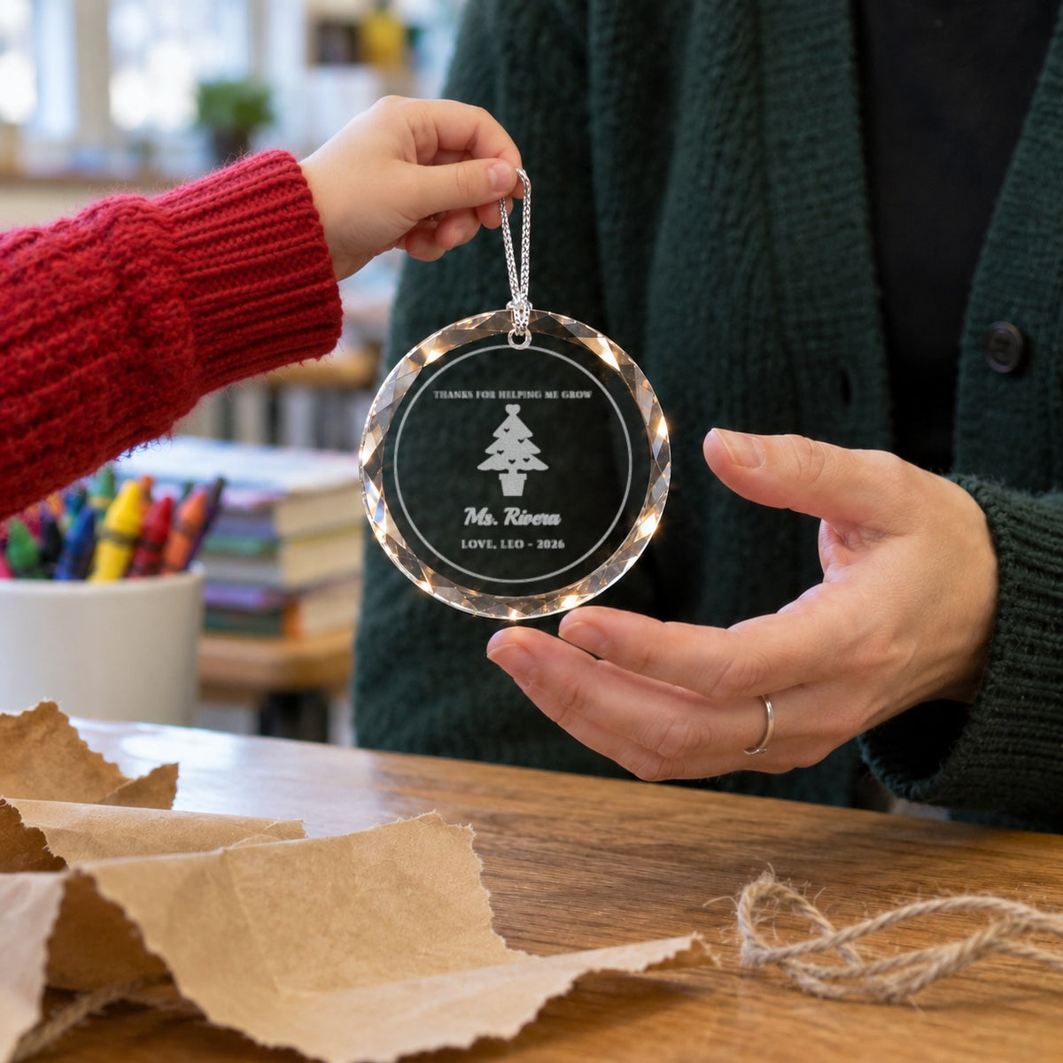 A child hands a personalized teacher ornament to her teacher over torn kraft paper, engraved THANKS FOR HELPING ME GROW for Ms. Rivera, LOVE, LEO 2026