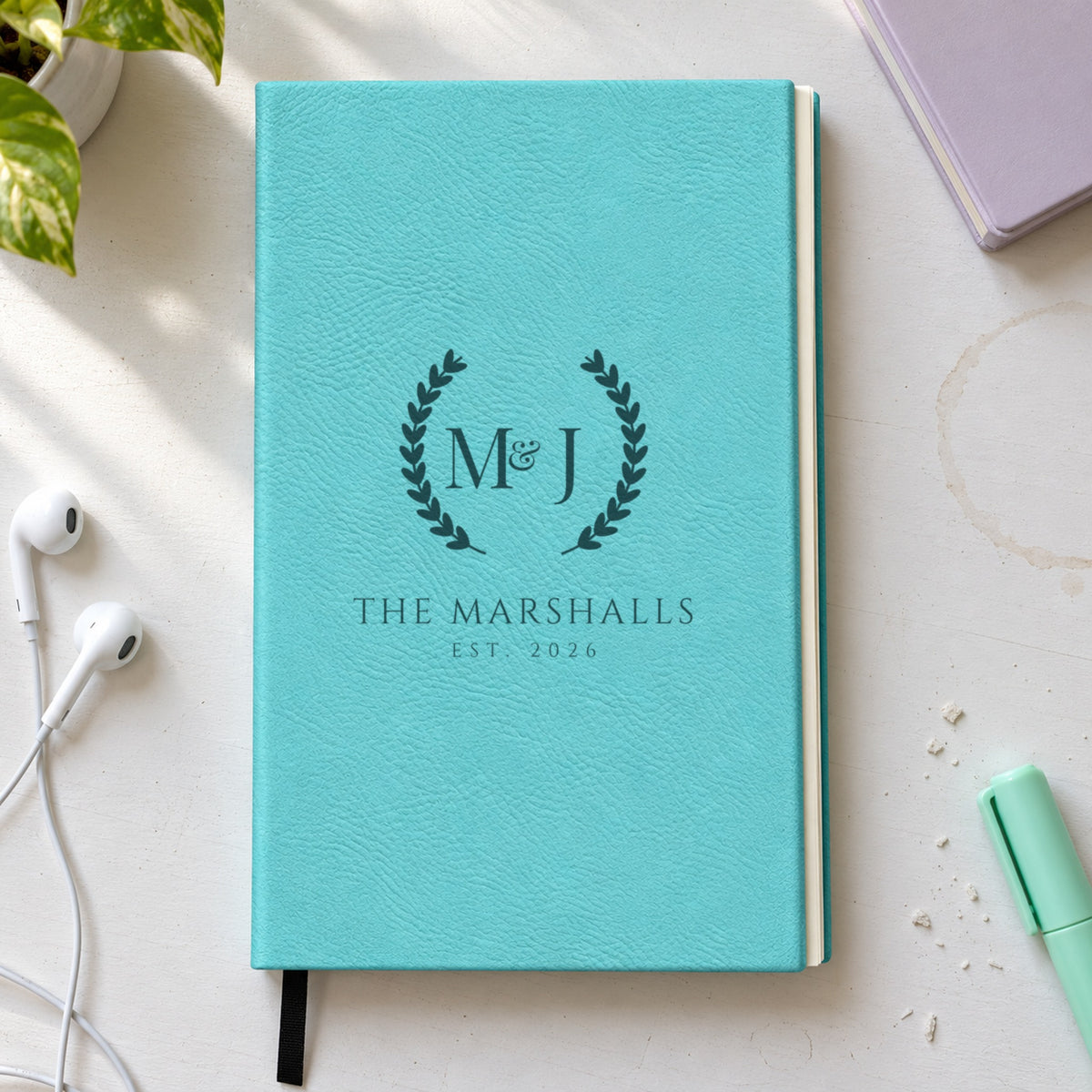 Teal personalized journal engraved with a wedding monogram, the initials M and J in a laurel wreath over The Marshalls and Est. 2026, on a white desk