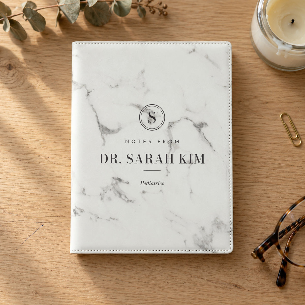 White marble personalized padfolio engraved Notes from Dr. Sarah Kim, Pediatrics, on an oak desk with reading glasses and a candle